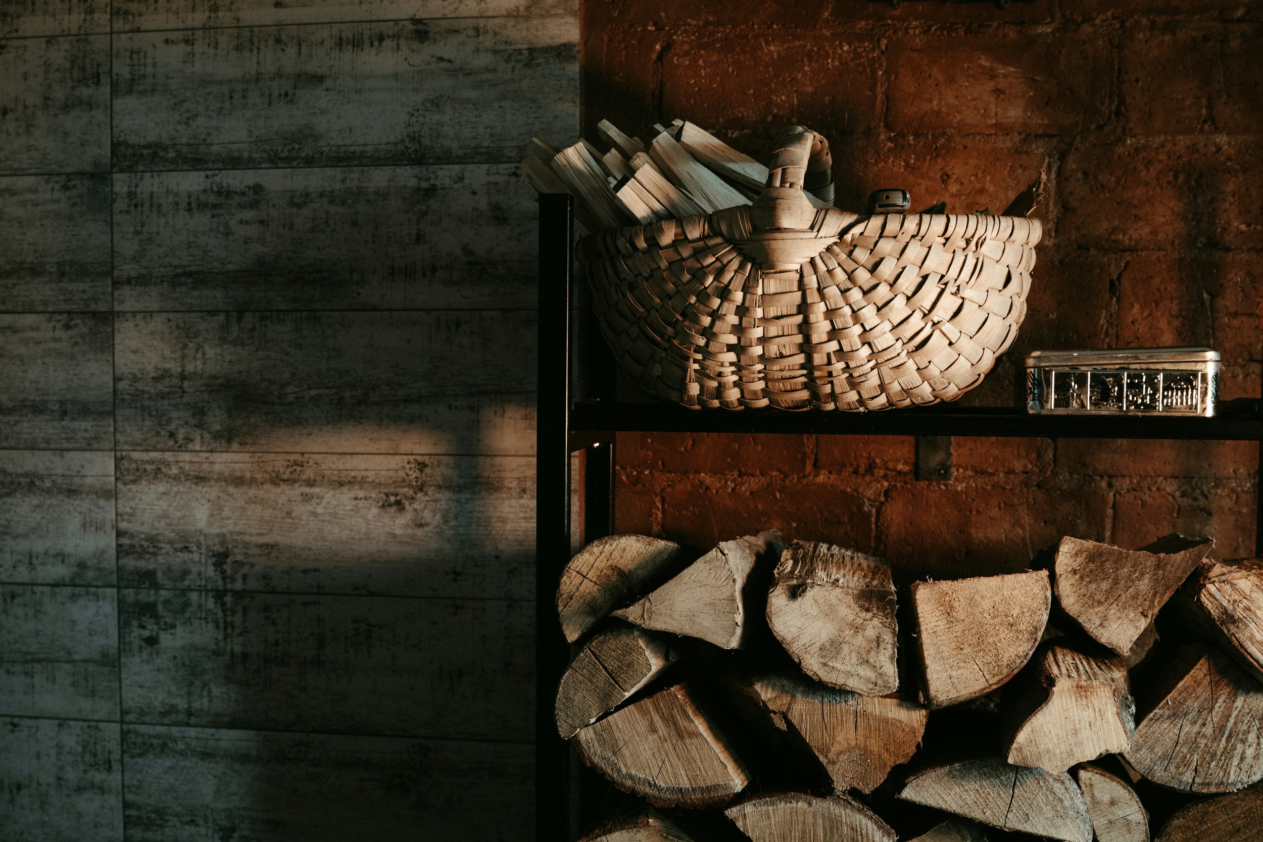Wood storage shelf with chopped firewood on the bottom and a woven basket with firewood and a small metallic container on top, against a wall with brick and wood paneling.
