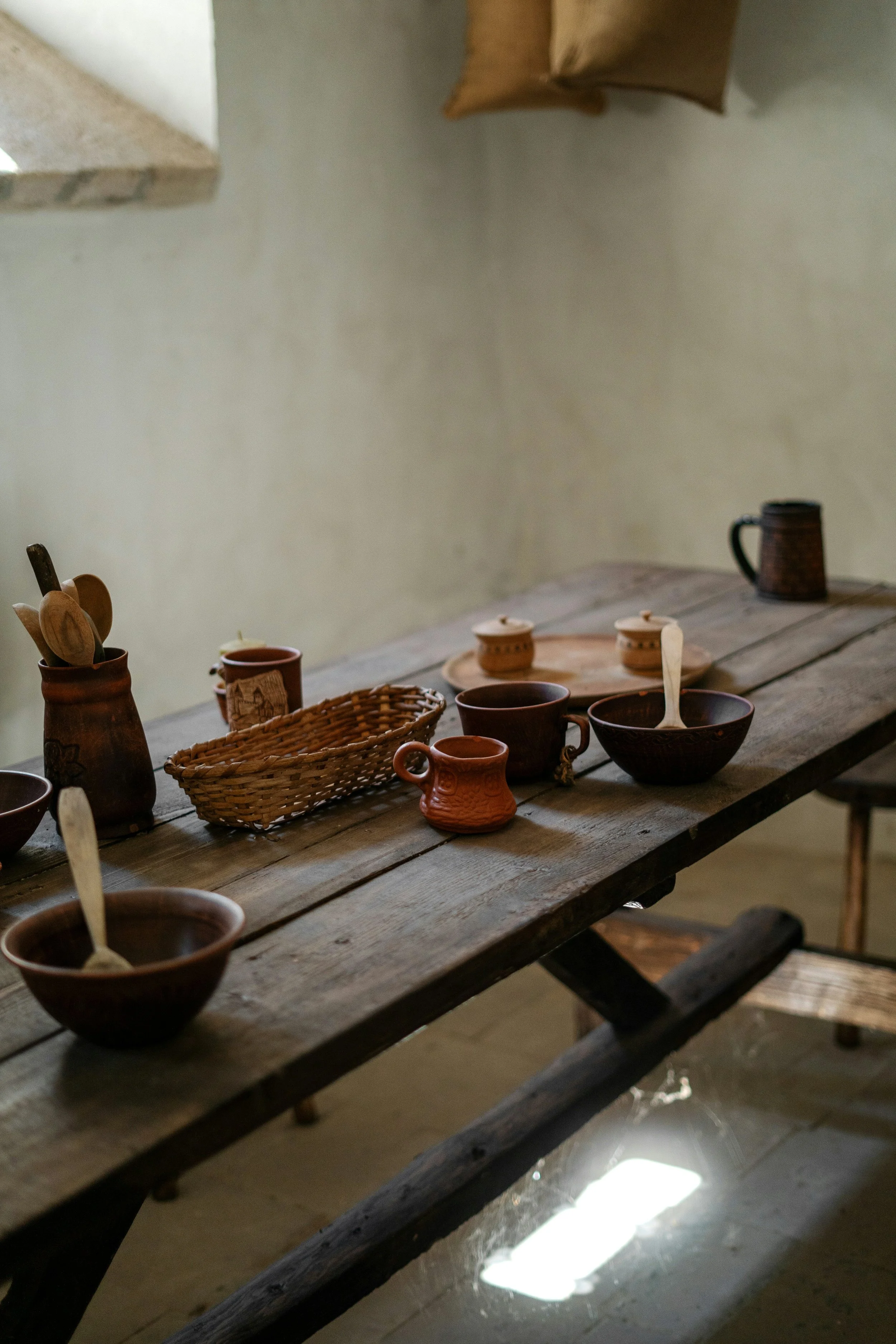 A rustic wooden table set with clay and wooden cups, bowls, and utensils, with a small basket, in a cozy, rustic setting.