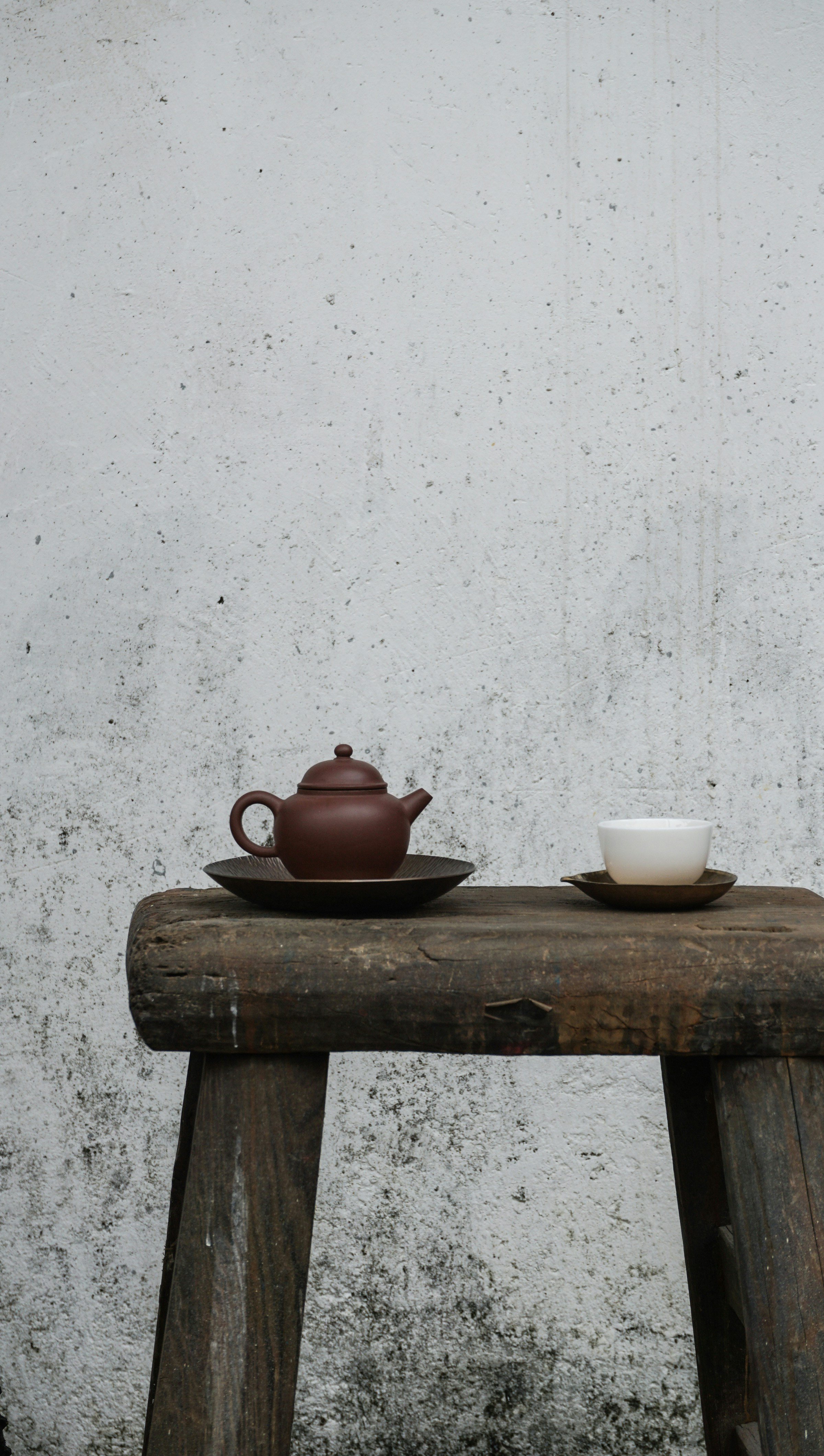 A small brown teapot and an empty white teacup on dark plates, placed on a rustic wooden table against a white textured wall.