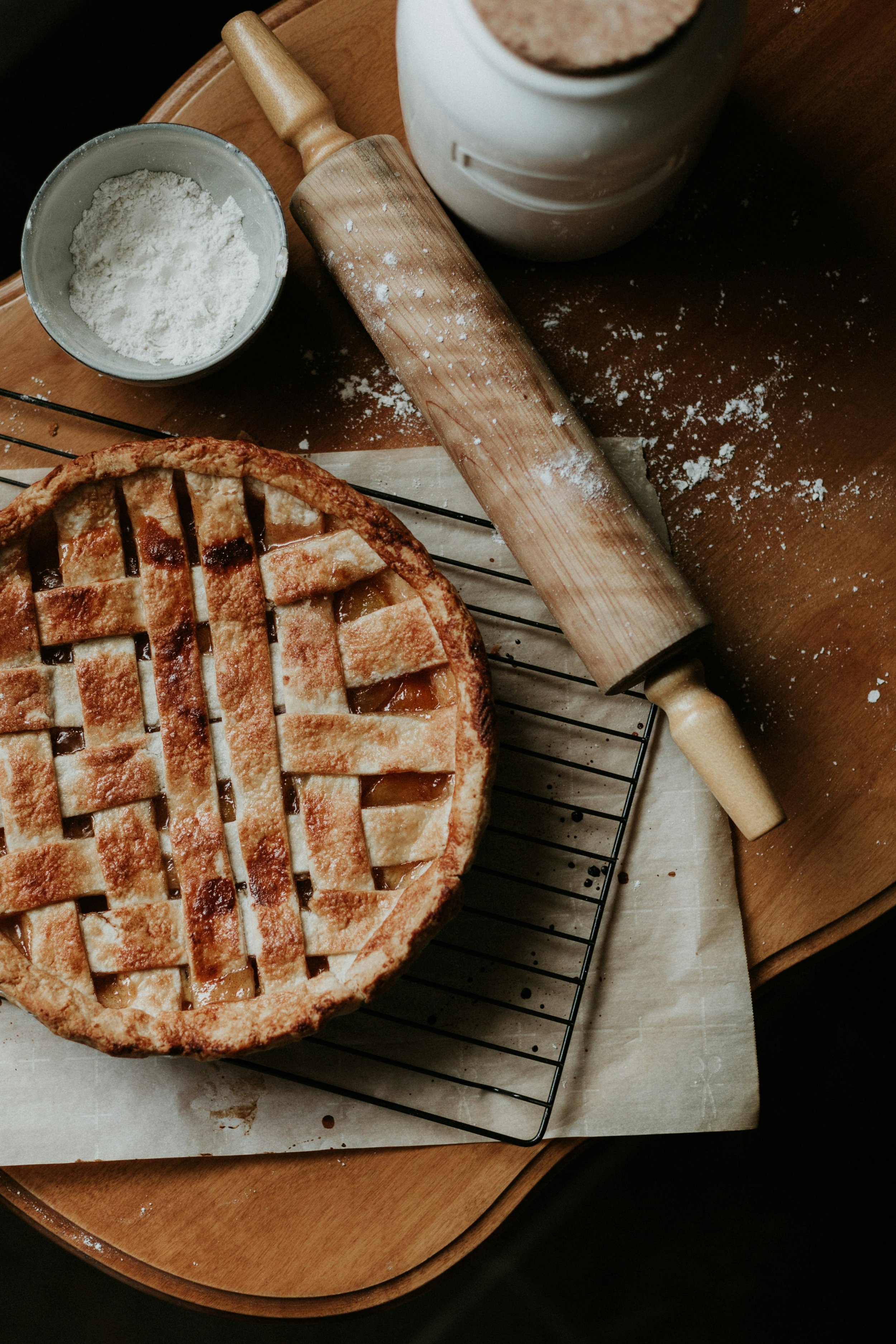 A freshly baked apple pie with a lattice crust on a cooling rack, surrounded by a rolling pin, a bowl of flour, and a jar of cinnamon, on a wooden table.
