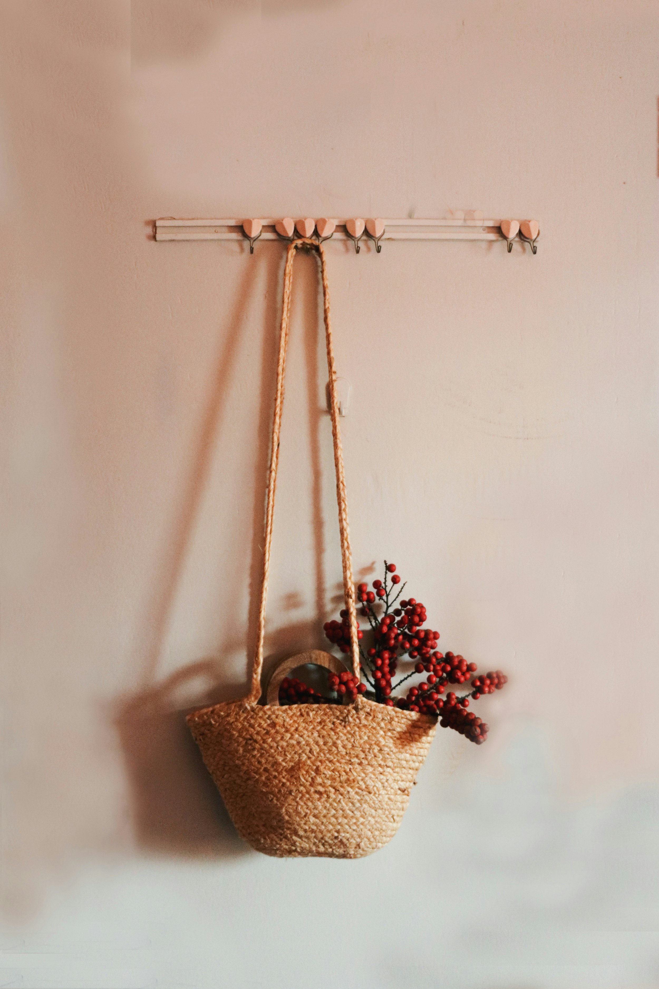 A woven straw bag hanging on a wall hook with red berry branches inside the bag, and a plain light-colored wall in the background.