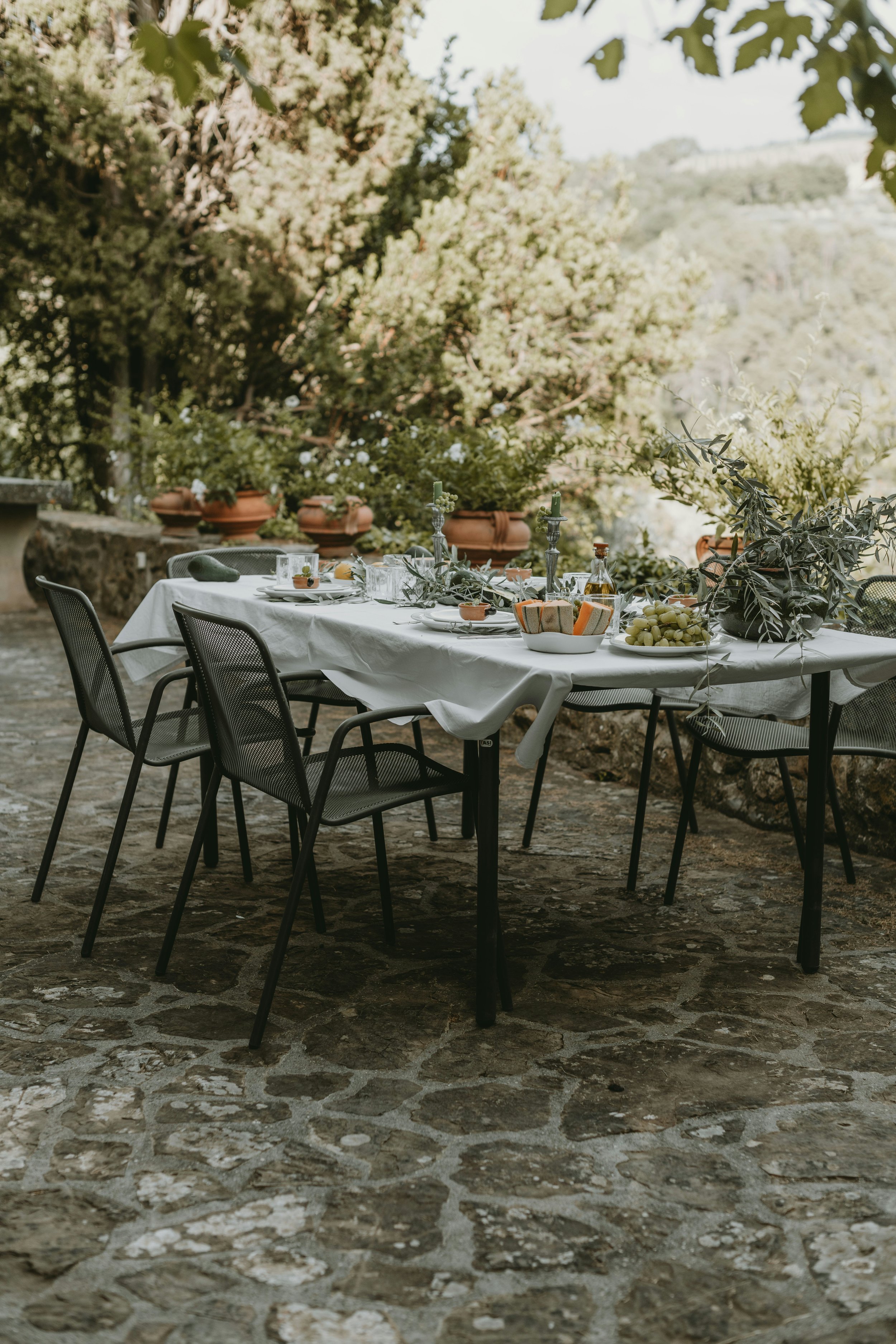 An outdoor dining table set with white tablecloth, surrounded by black chairs, on a stone patio with potted plants and greenery in the background.