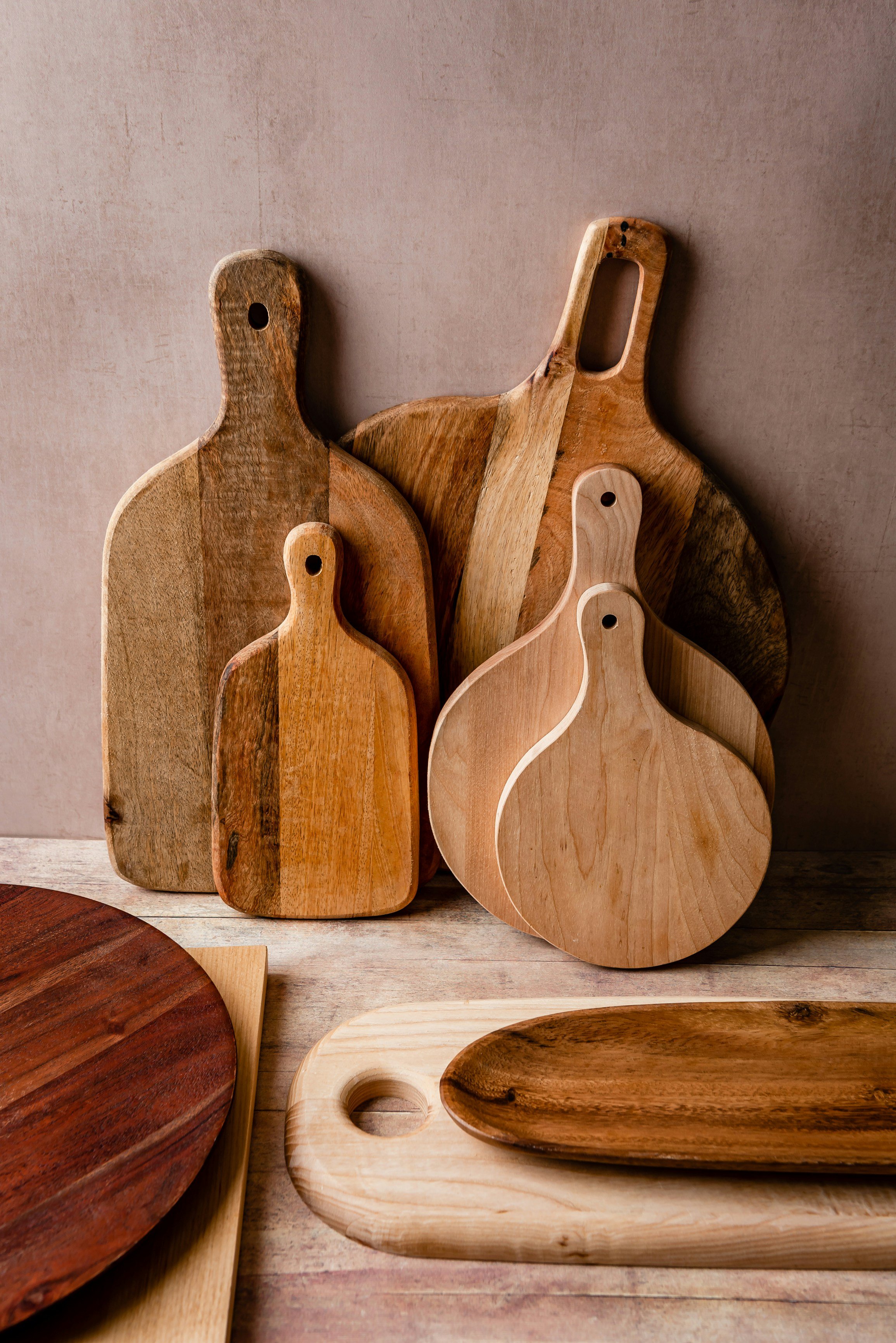 Collection of wooden cutting and serving boards of various shapes and sizes, displayed on a wooden surface against a pinkish wall.