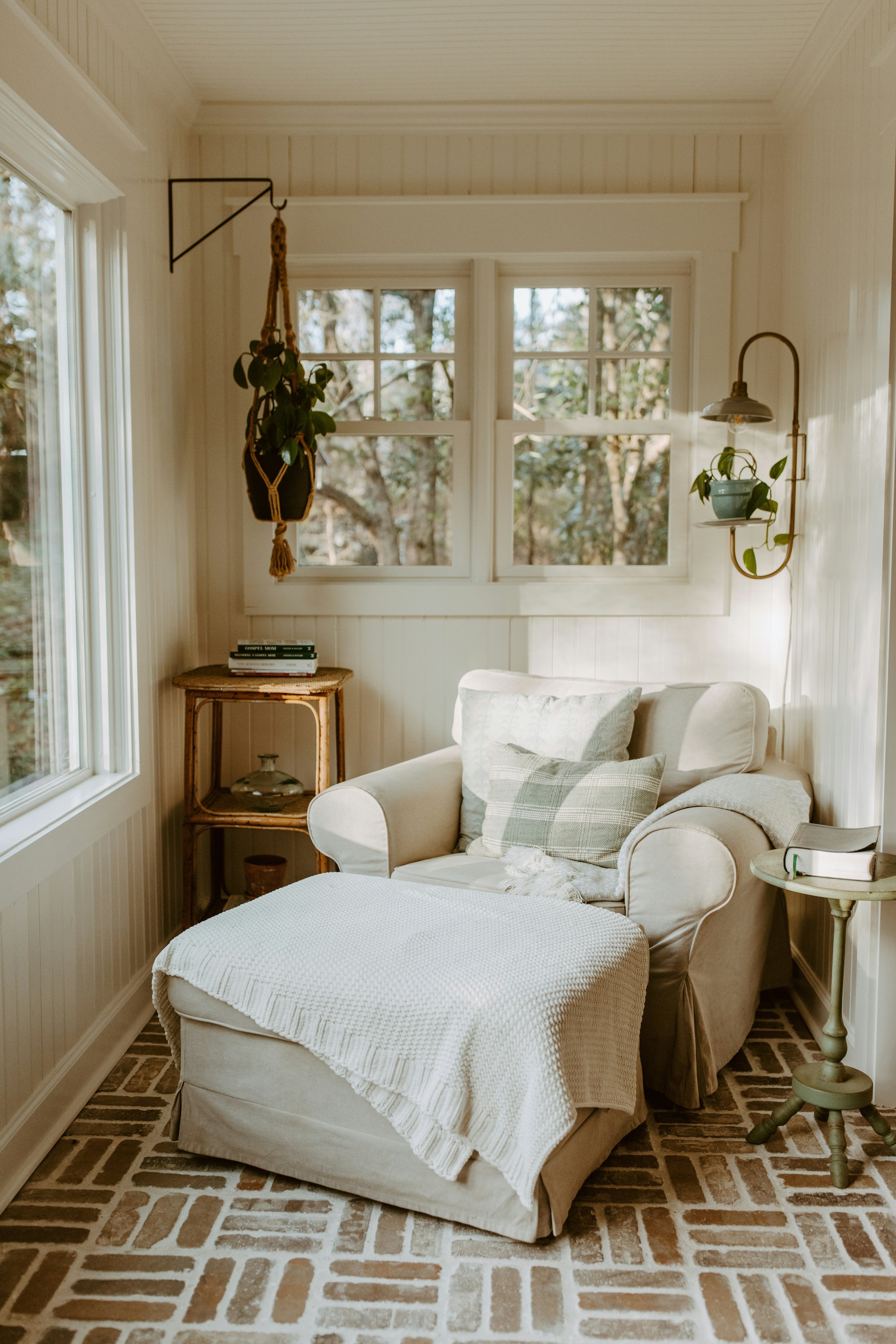 Sunlit cozy reading nook with beige armchair, white throw blanket, patterned pillows, side table with books, brick floor, window views of trees, hanging plant, wall-mounted lamp with potted plant.