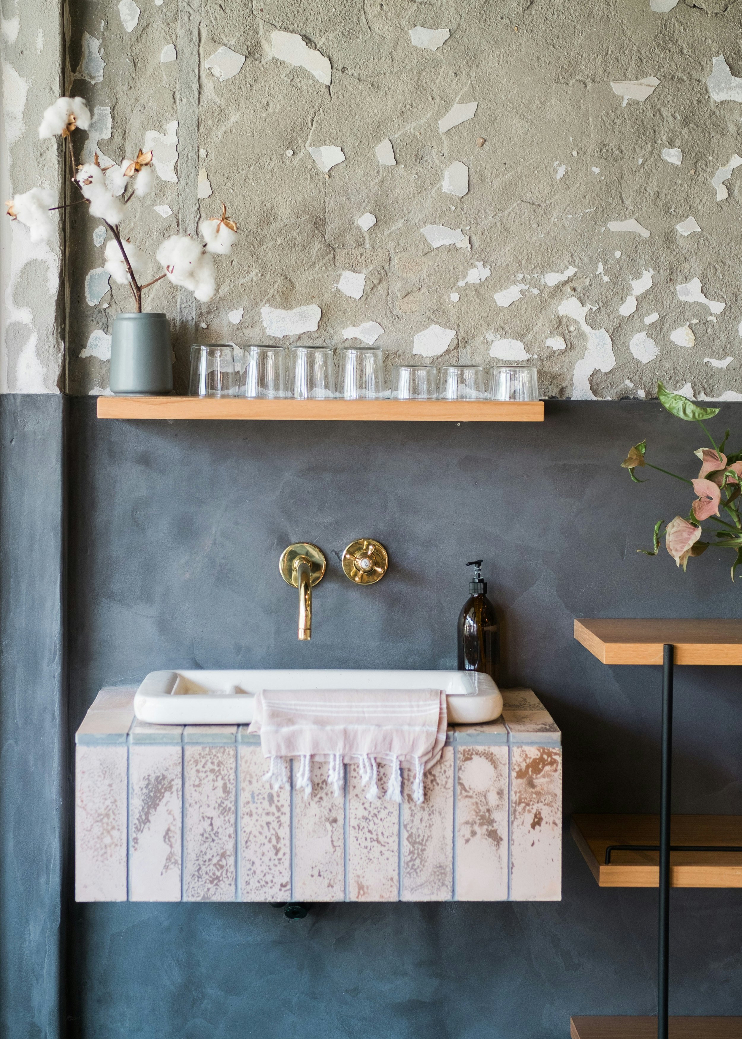 Interior of a rustic bathroom with a concrete wall, a tiled sink with a towel, a black soap dispenser, a wooden shelf with glasses and a vase with cotton branches, and a plant on a nearby wooden shelf.