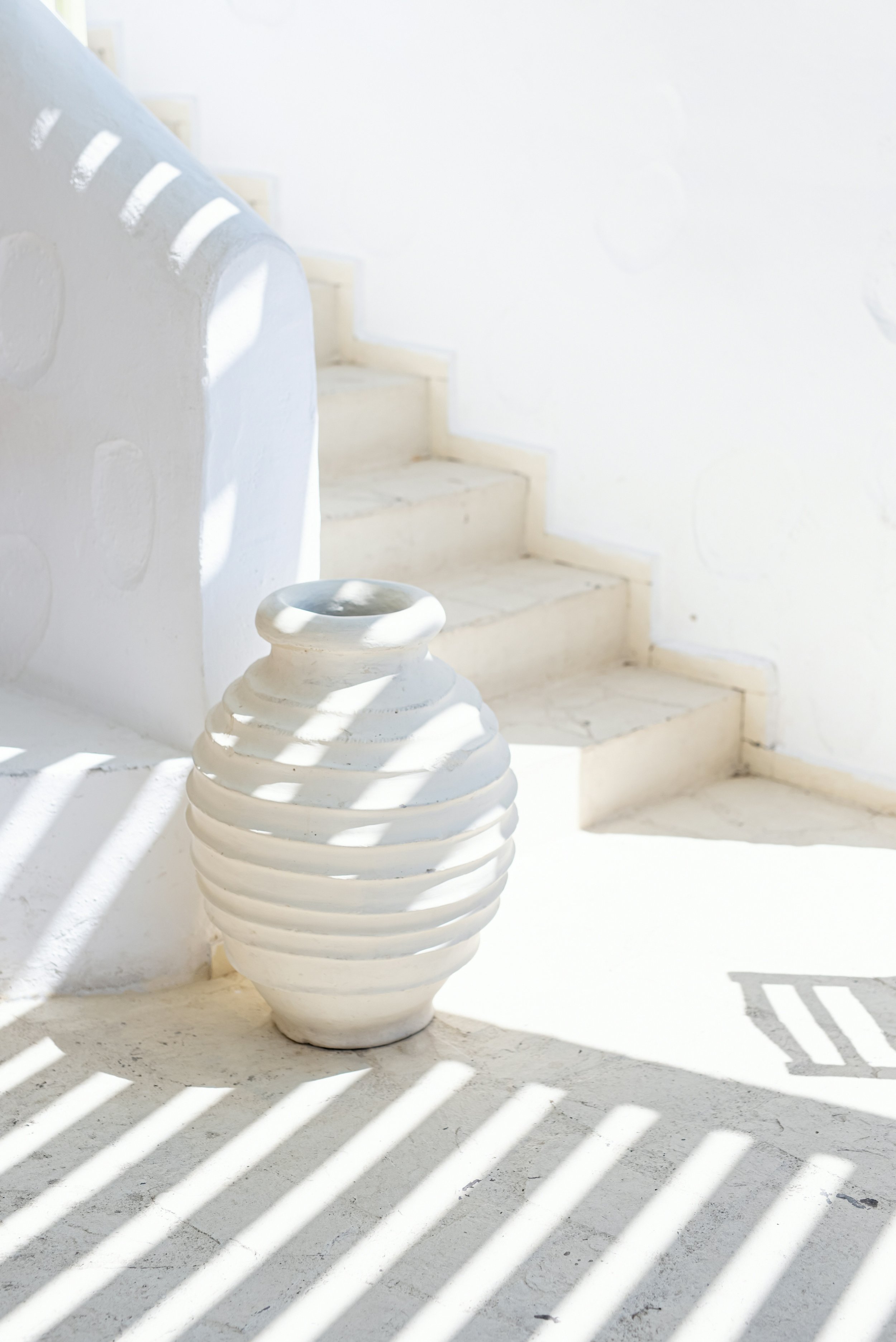 White ceramic vase with horizontal ridges near white stairs and a white wall, sunlight casting striped shadows.