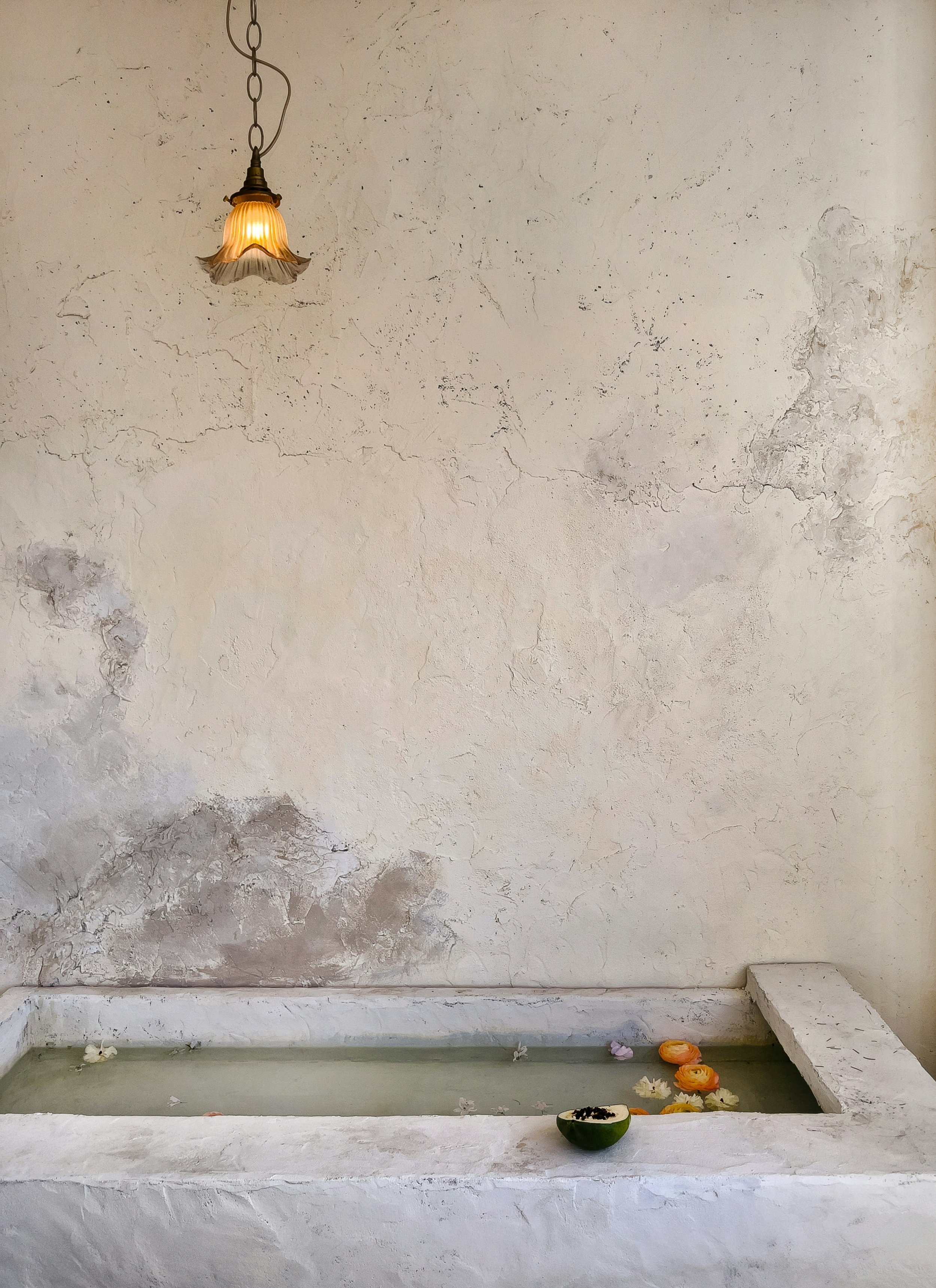 A rustic indoor fountain with water and floating flowers, a small bowl on the edge, a vintage hanging lamp on a distressed white wall.