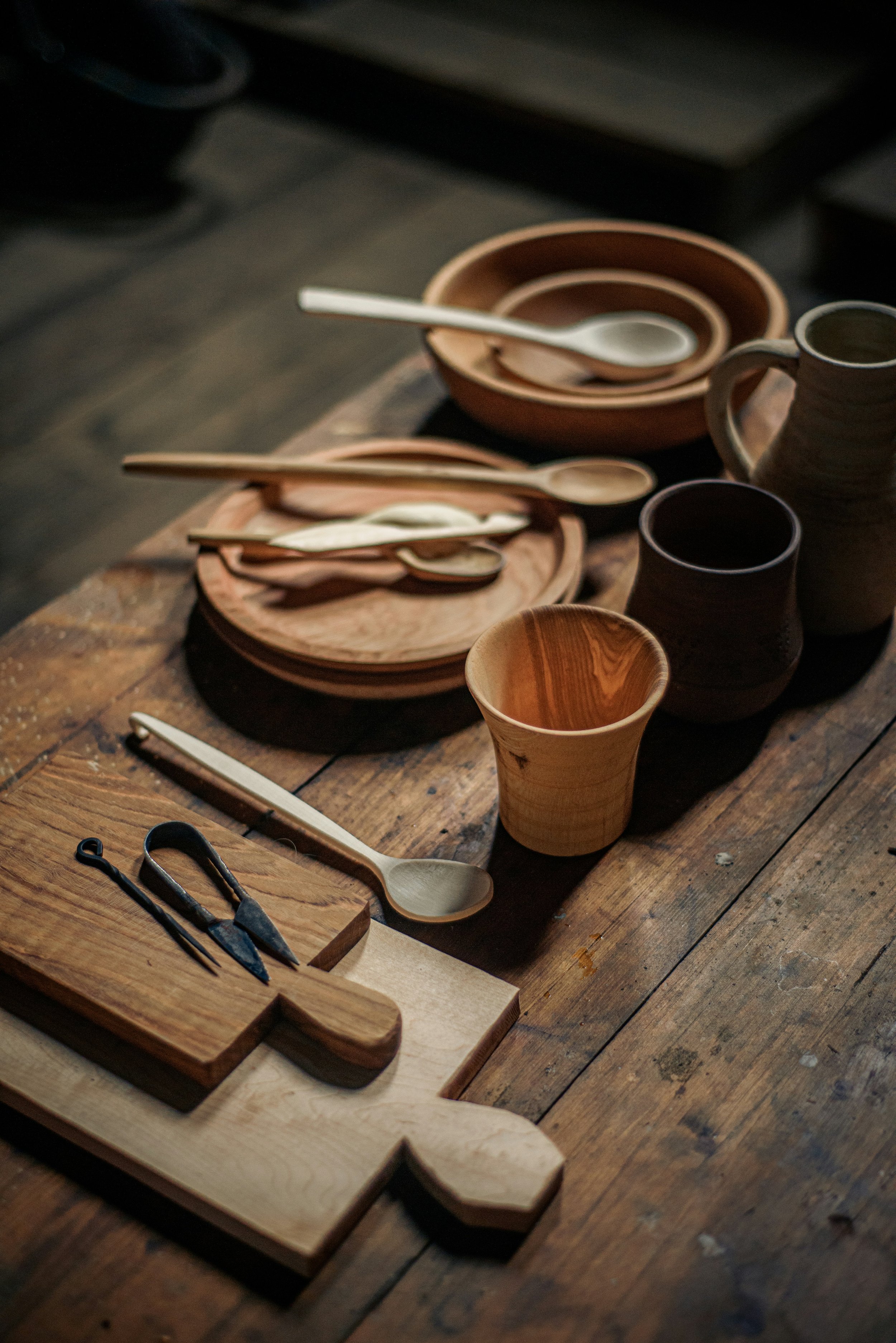 Various handmade pottery cups, bowls, and spoons arranged on a rustic wooden table, with small woodworking tools in the foreground.