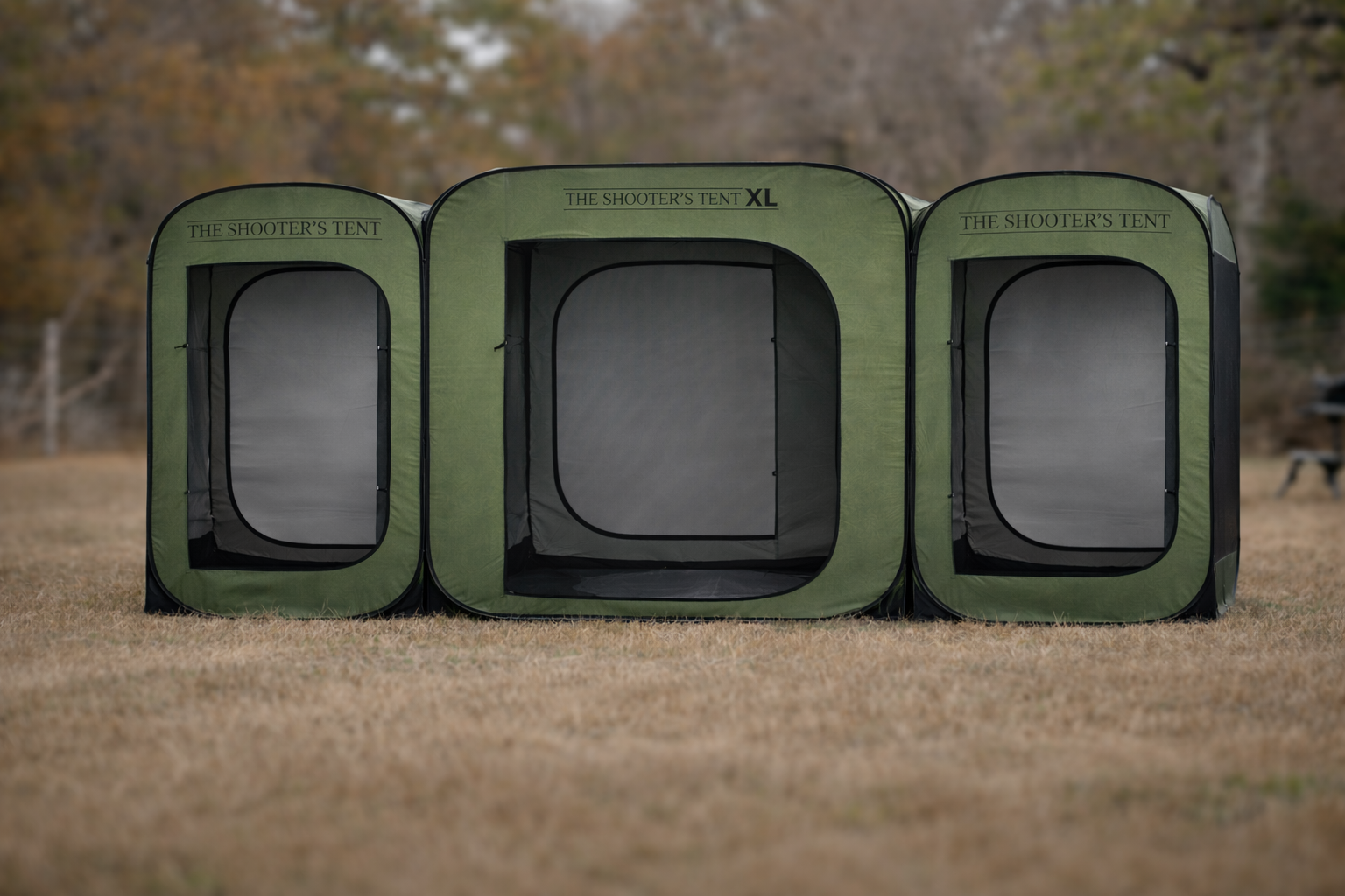 Green outdoor tent labeled 'The Shooter's Tent XL' set up on grass with trees in the background.