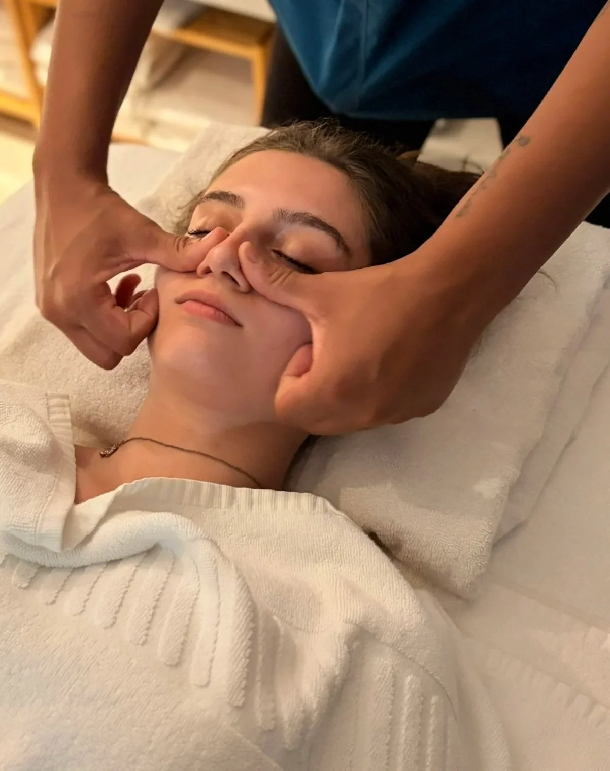 Pessoa recebendo uma massagem facial relaxante, com olhos fechados, deitada de costas em uma cama ou massageador, com alguém aplicando pressão na nariz com as mãos.
