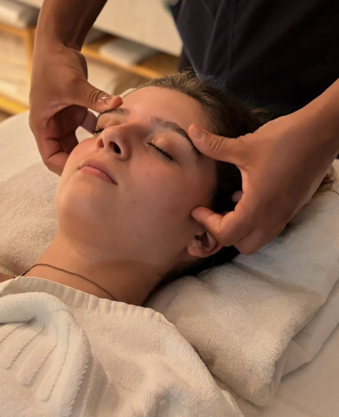 Pessoa recebendo uma sessão de terapia de face, deitada com olhos fechados, enquanto um profissional faz massagem facial na testa e nas bochechas.