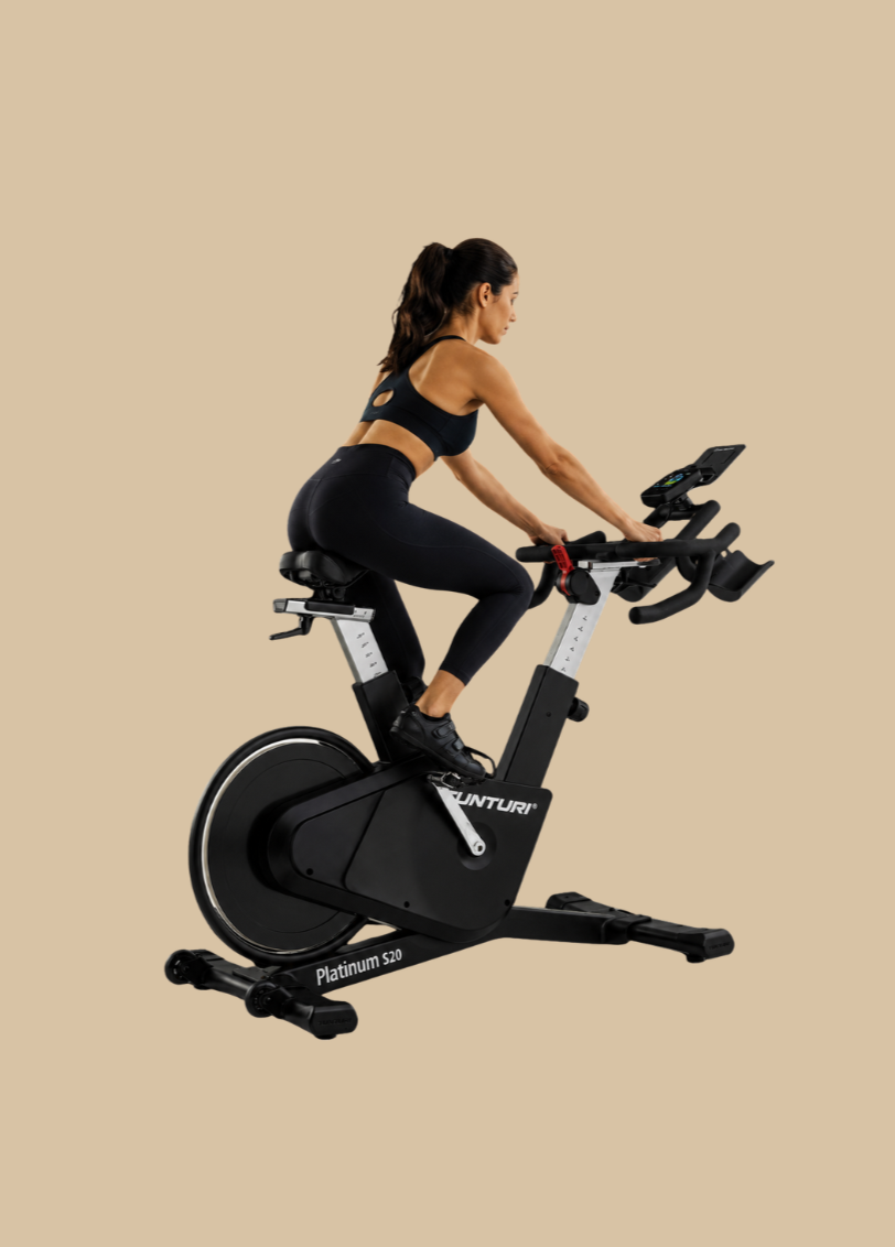 Mulher treinando em bicicleta ergométrica, vestida de roupa preta, em fundo bege.