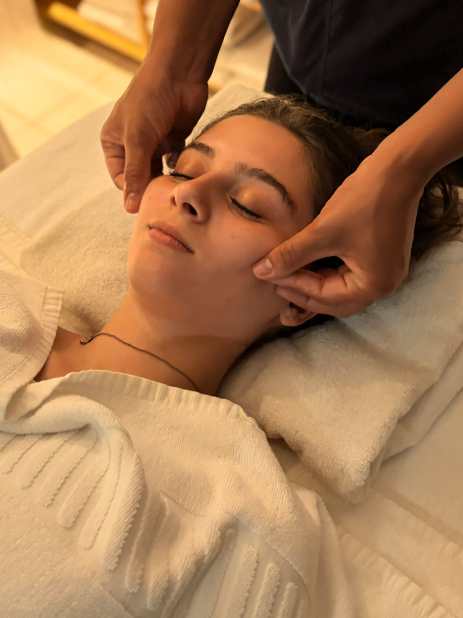 Pessoa recebendo uma massagem relaxante na cabeça enquanto descansa em uma cama com roupas brancas, com uma toalha branca ao lado.