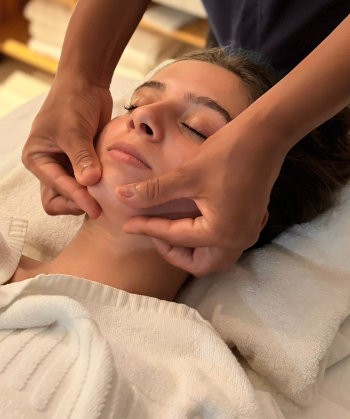 Pessoa recebendo uma massagem relaxante no rosto, deitada de costas com olhos fechados, enquanto alguém realiza a massagem com as mãos.