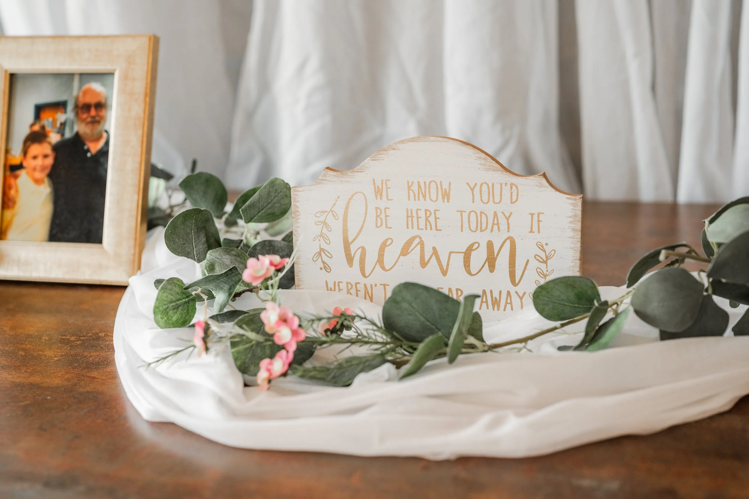 Wedding memorial table sign surrounded by greenery, soft fabric, and personal photos as a meaningful remembrance display