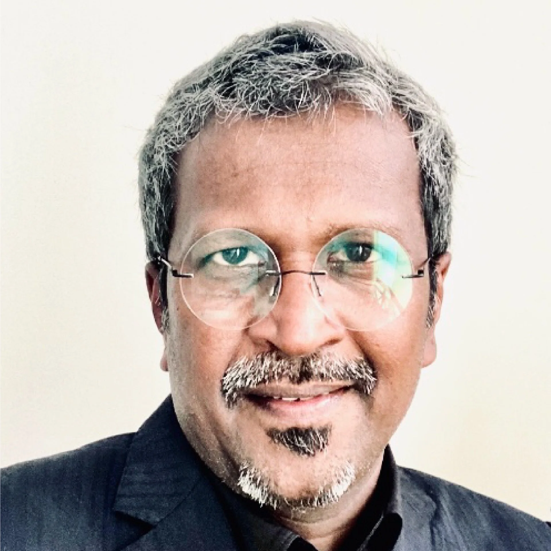 A man with salt-and-pepper hair and beard wearing round glasses and a dark suit, smiling against a plain white background.