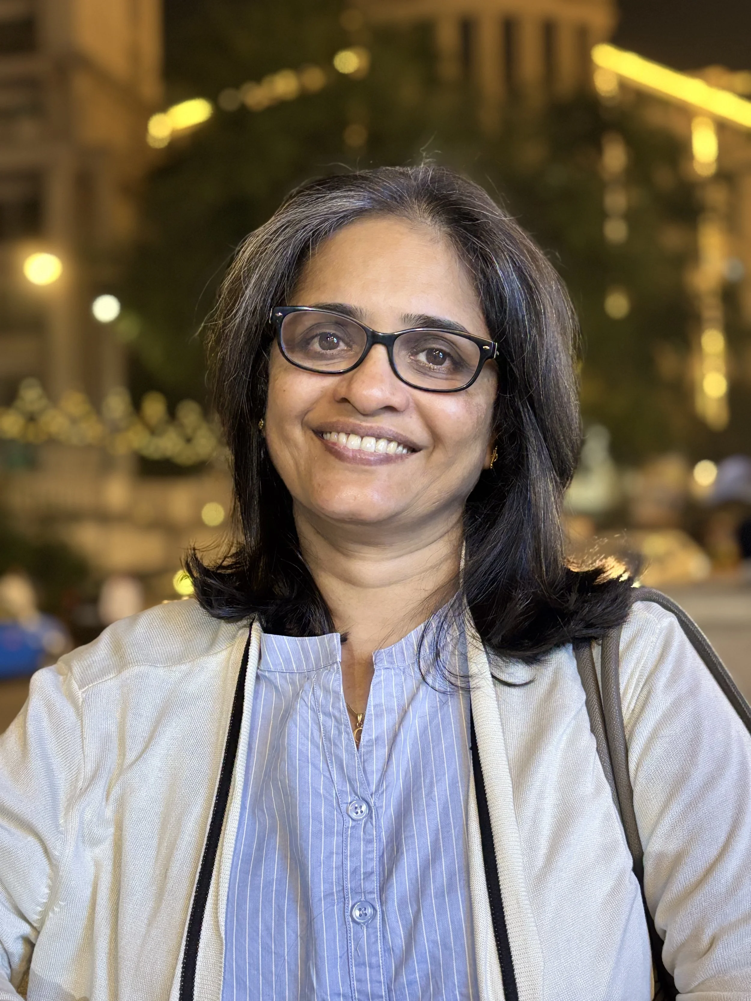 Smiling woman wearing glasses, blue striped shirt, and light-colored jacket, outdoors at night with blurred city lights and buildings in the background.