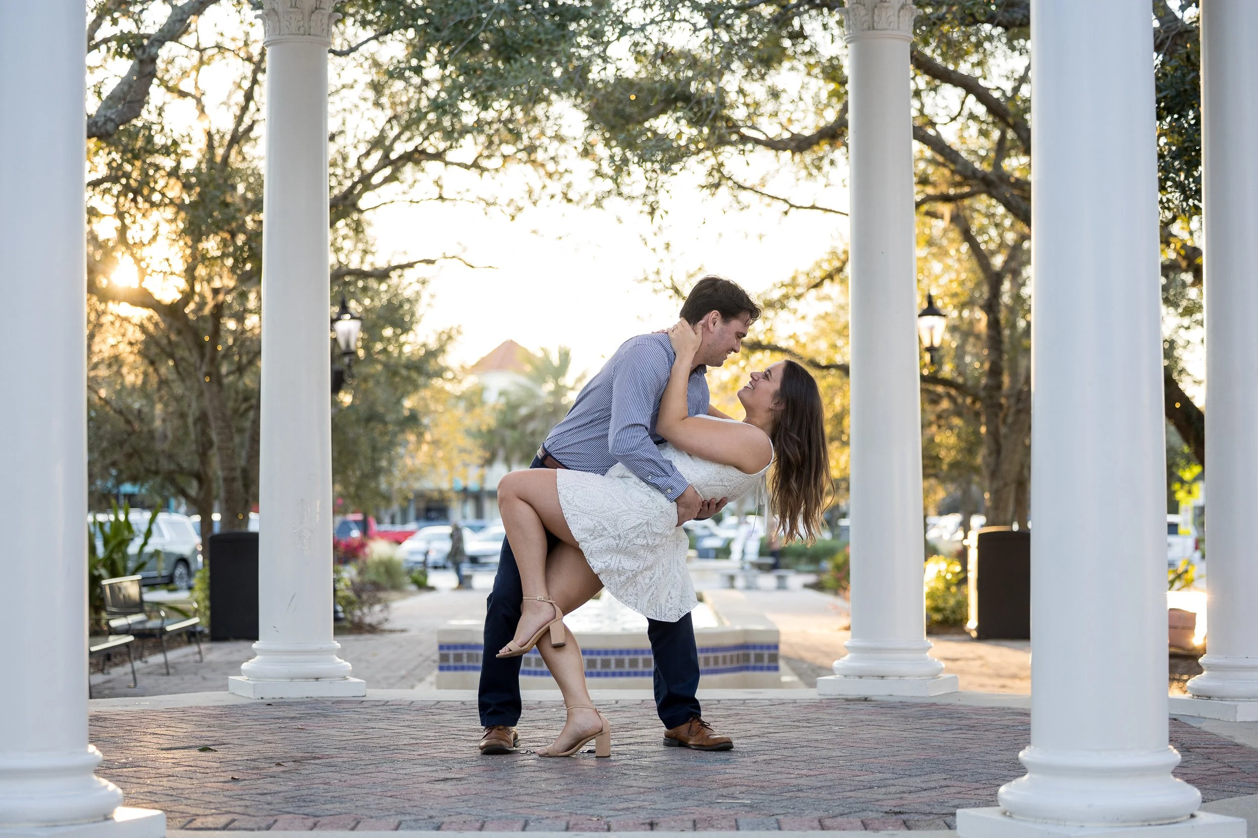 A couple posing under a pergola, with the man dipping the woman playfully. The woman is wearing a white dress, and the man is wearing a blue shirt and dark pants. The background features trees and a sunset glow.