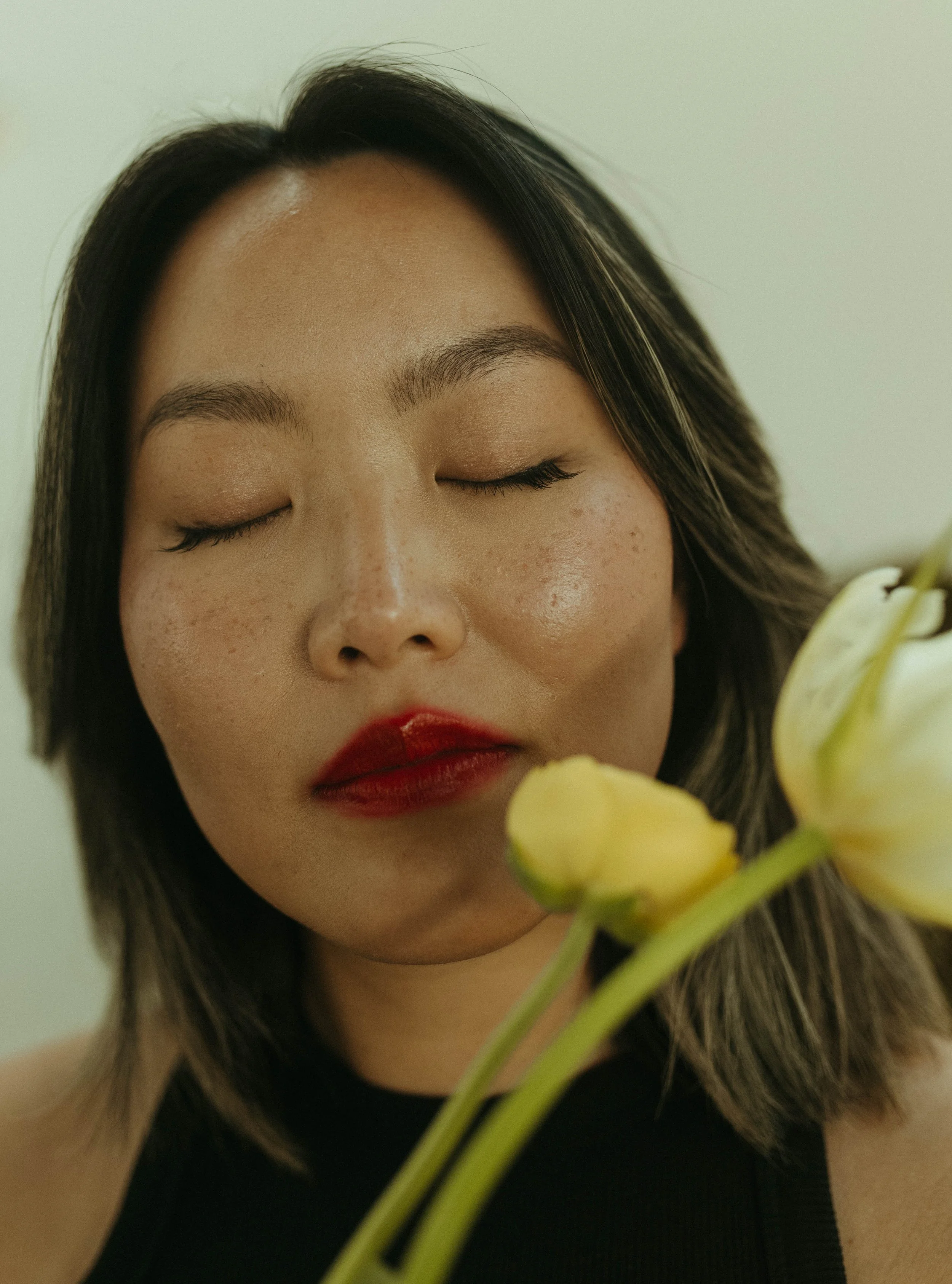 Close-up of a woman with closed eyes, dark hair, and red lipstick, holding a yellow flower near her face.
