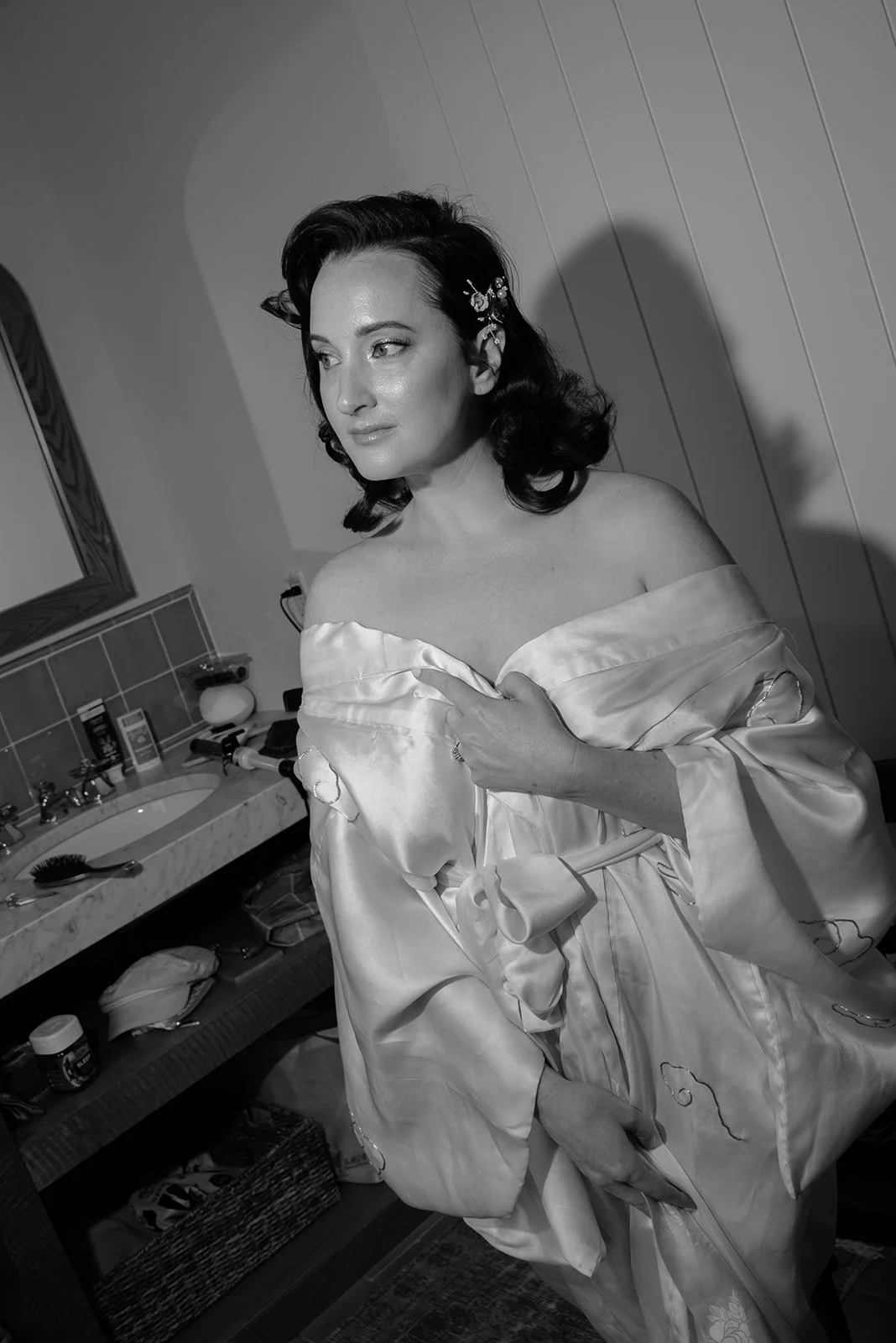 A woman with dark hair styled in waves, wearing a silky, off-shoulder robe, adjusting it with her right hand while looking to the left in a bathroom. The bathroom countertop has various items, including bottles and personal care products.