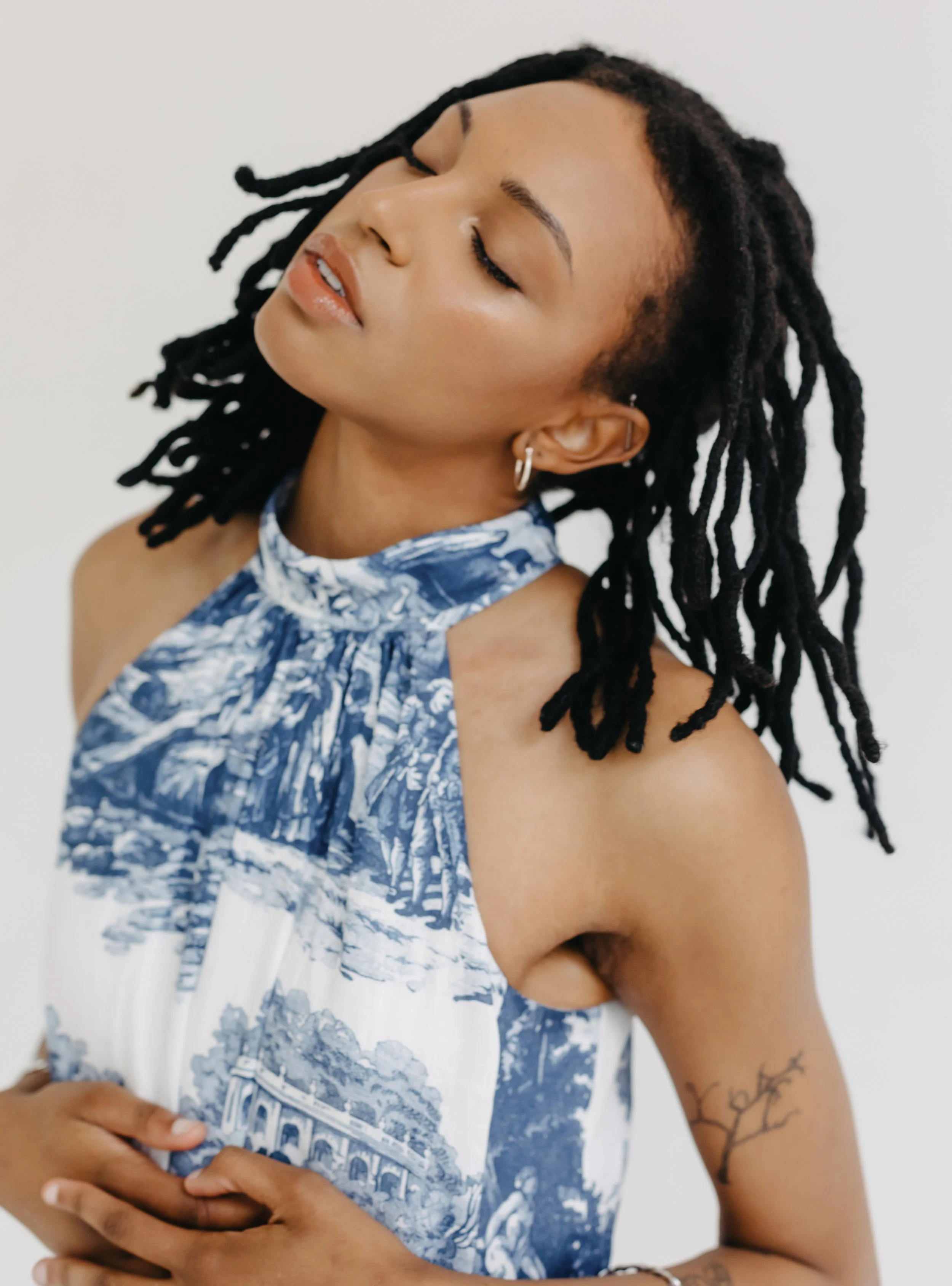 A woman with long dreadlocks, wearing a sleeveless blue and white patterned top, with her eyes closed and one hand resting on her stomach.