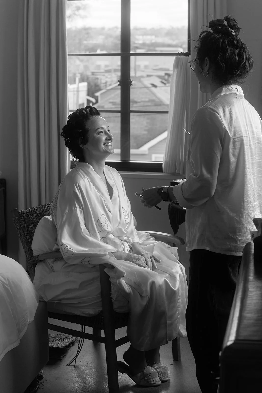 A woman in a chair wearing a satin robe smiling at a stylist or makeup artist standing beside her, in a room with a large window in the background.