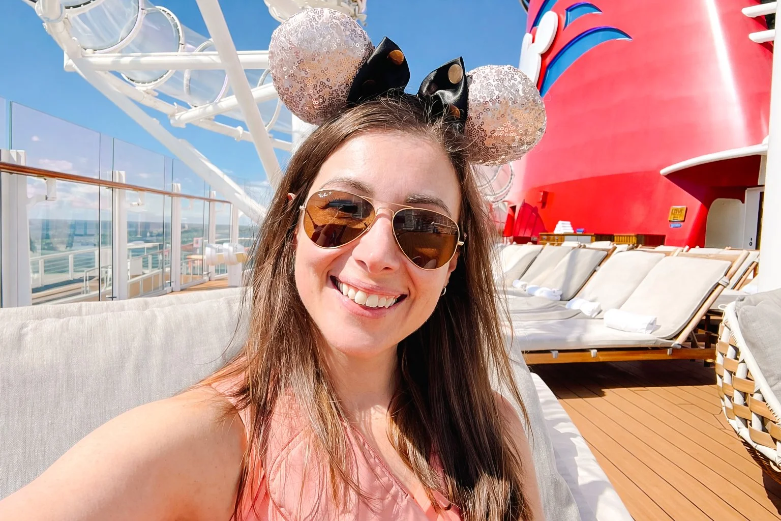 A woman with brown hair wearing sunglasses and Disney Minnie Mouse ears hat, smiling on the deck of a cruise ship with lounge chairs and a large red structure in the background.