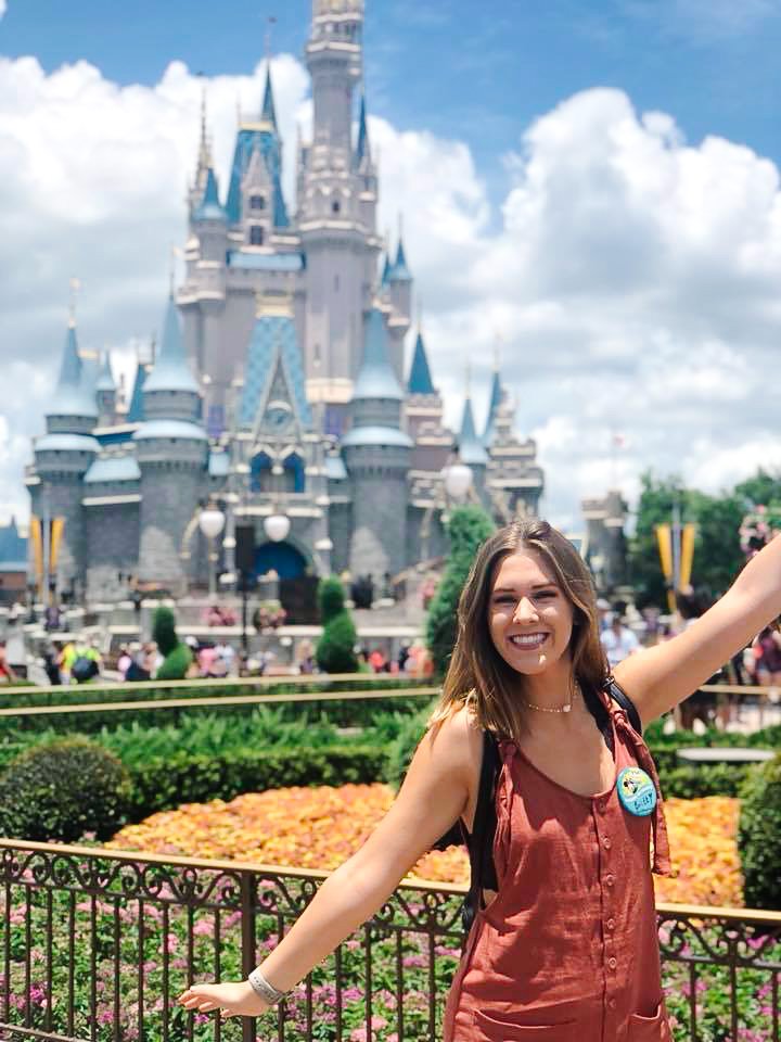 A young woman with a backpack and a button on her shirt is smiling and posing with her arms outstretched in front of Sleeping Beauty Castle at Disneyland during daytime. She is wearing a sleeveless, rust-colored dress and standing in a landscaped garden with colorful flowers.