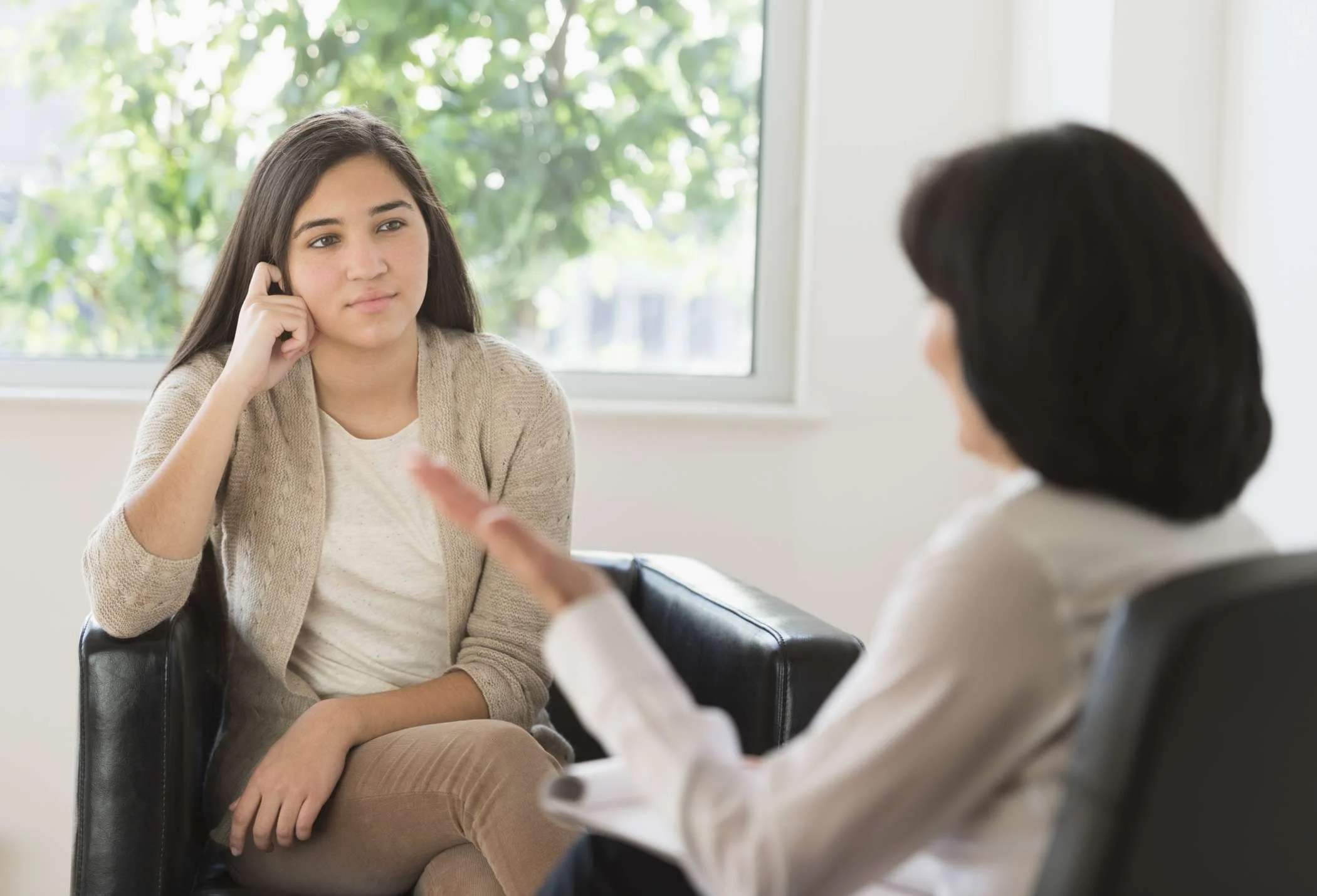 A young woman in a beige cardigan and white shirt sitting and listening during a therapy or counseling session, talking to a therapist with short dark hair, wearing a white top, inside a room with a window showing greenery outside.