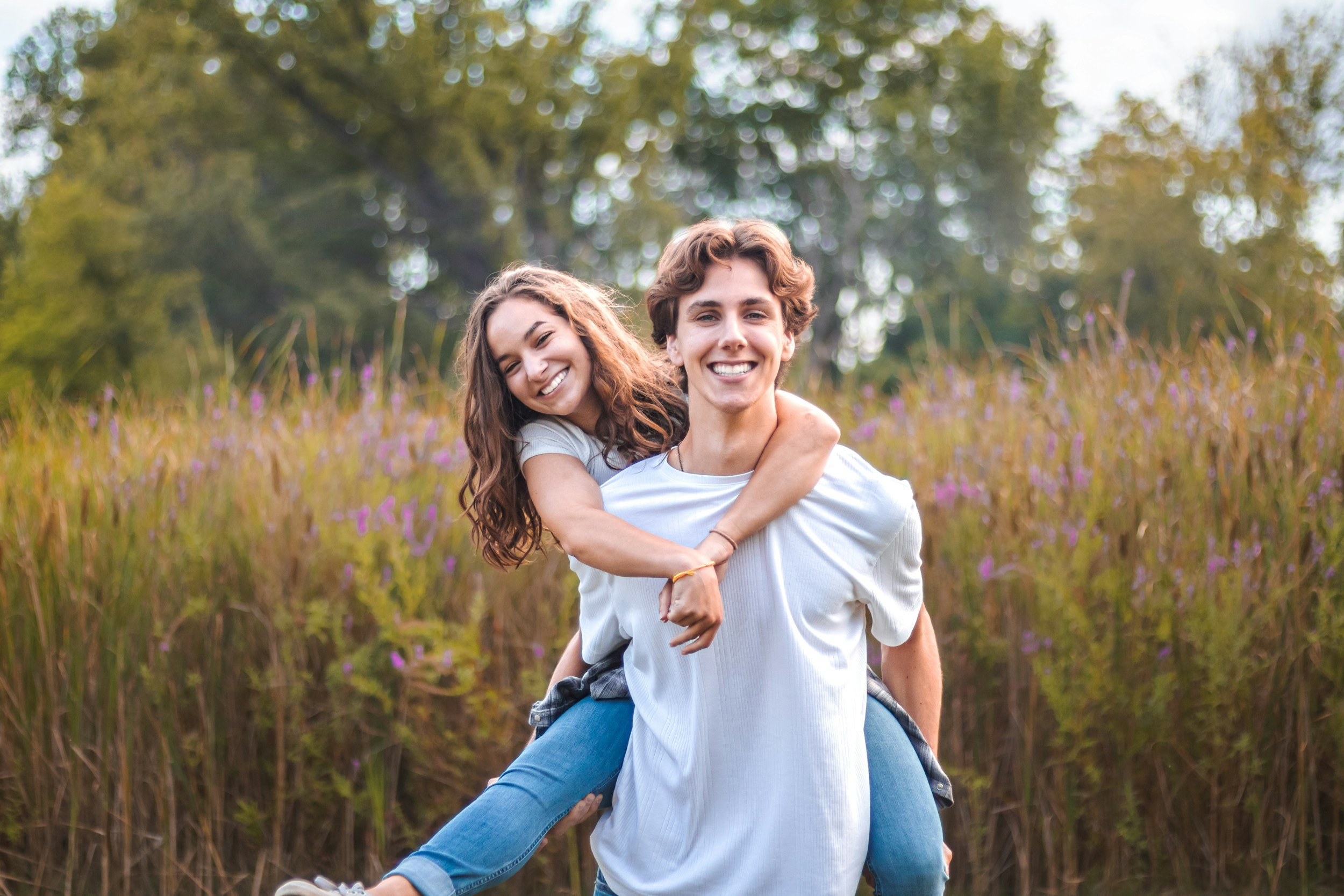 A young man giving a piggyback ride to a smiling young woman in a field with tall purple wildflowers and green trees in the background on a sunny day.