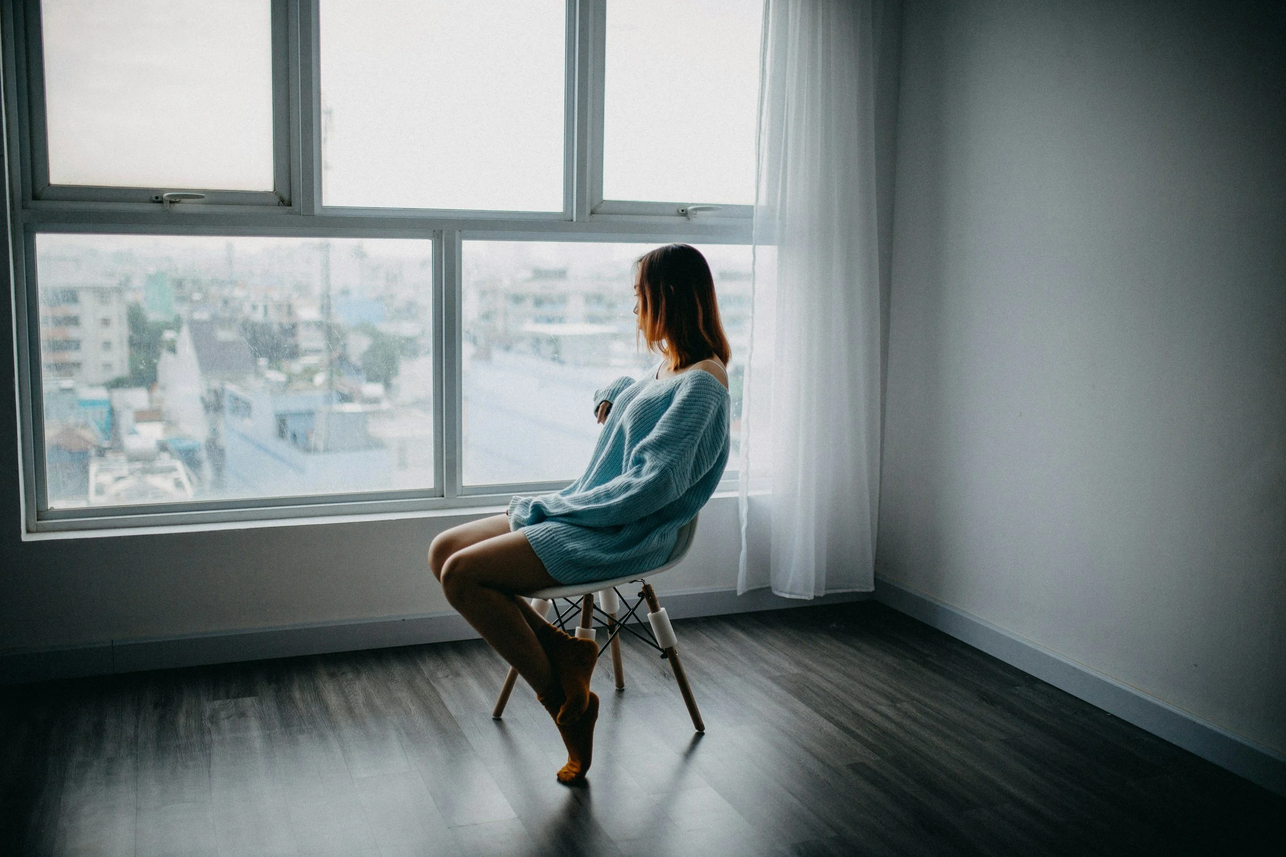 A woman sitting on a chair by a large window, looking outside at a cityscape, wearing an oversized blue sweater and brown socks.