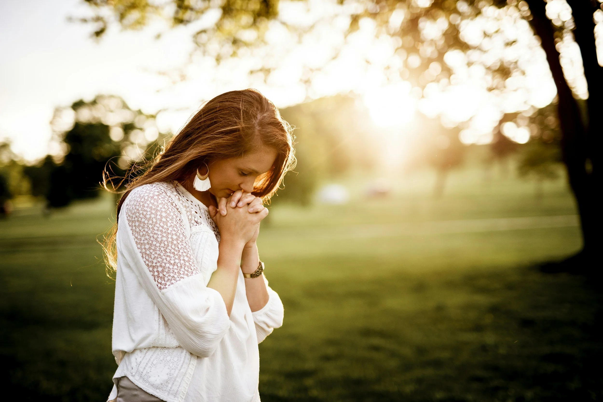 A woman with red hair, wearing earrings and a white blouse with lace details, is standing outdoors with her hands clasped and her head bowed in prayer or reflection as the sun sets behind her in a park with green grass and trees.
