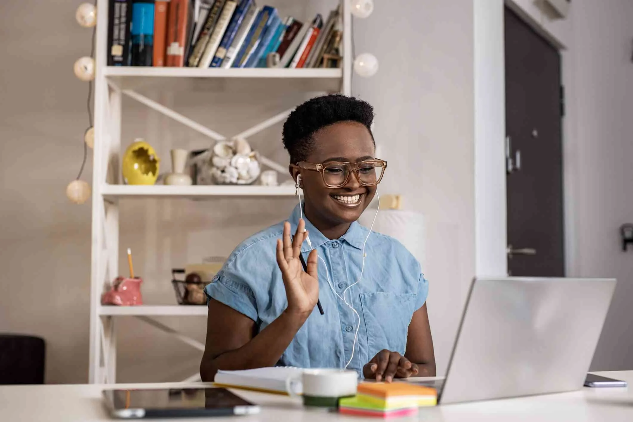 Smiling woman with short black hair and glasses participating in a video call on her laptop, waving, with a pen in her hand, sitting at a desk with notebooks, a smartphone, a mug, and a stack of sticky notes, in a room with a bookshelf and a closed door in the background.