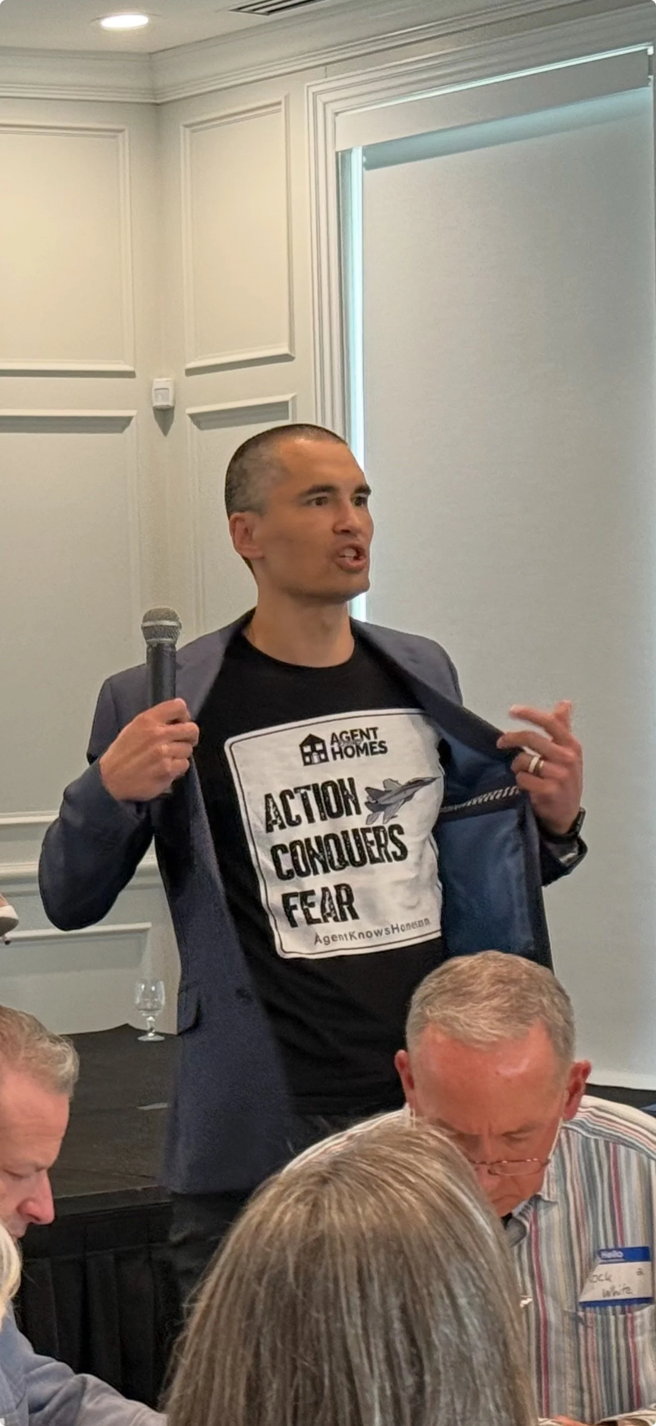 Image of Jeff Wu. A man speaking into a microphone is standing and wearing a T-shirt that reads 'Action Conquers Fear' under an open blazer, with others seated around him at what appears to be a conference or meeting.