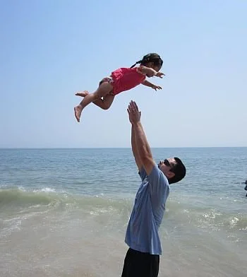 Image of Jeff Wu and his daughter. A man throwing a young girl into the air on a beach with the ocean and sky in the background.