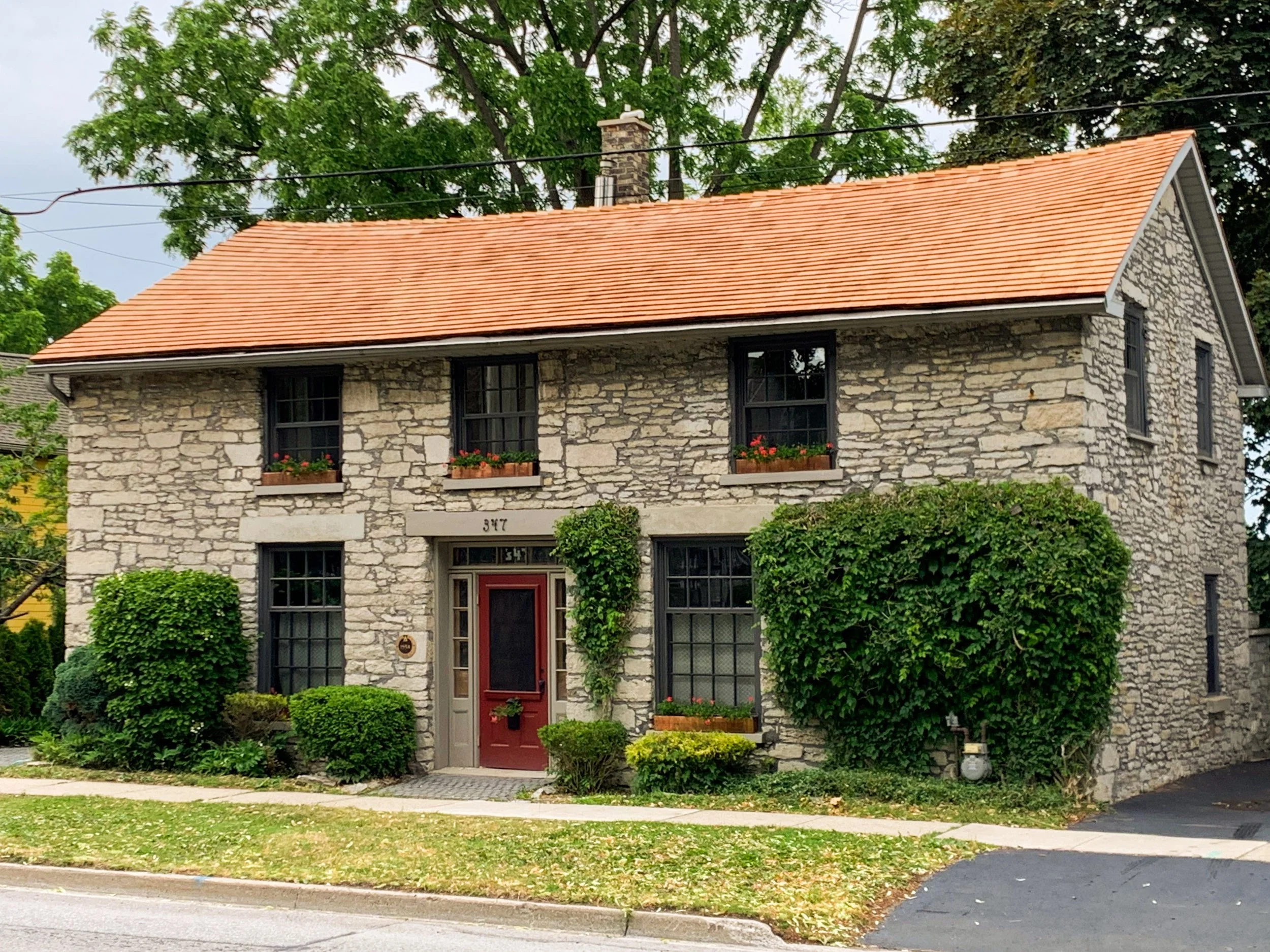 A two-story stone house with a red-tiled gable roof, black window frames, and a red front door, surrounded by green bushes and trees.