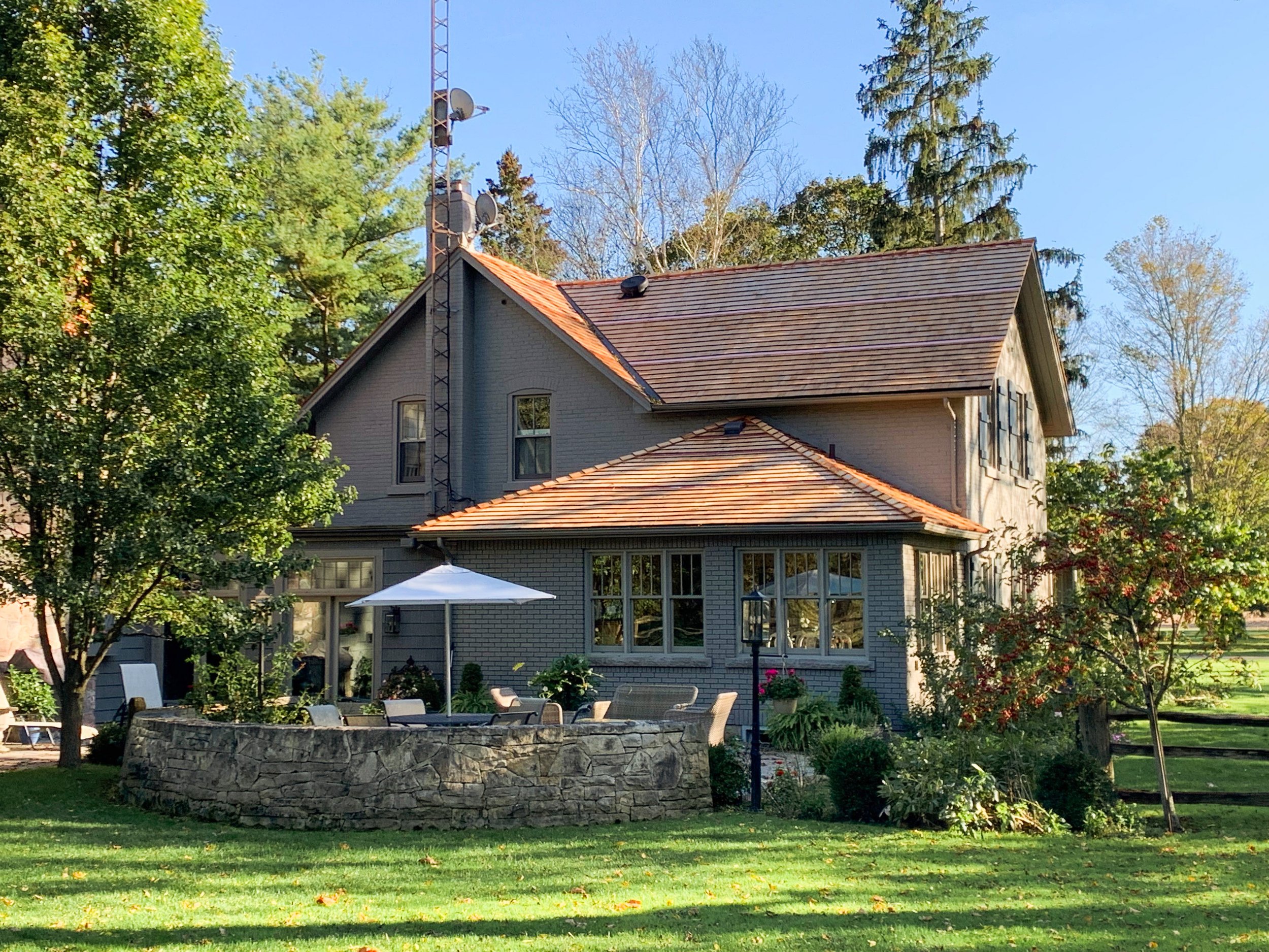 A two-story house painted light gray, with a steep gabled roof covered in red tiles, surrounded by a lush green lawn, mature trees, and a stone patio with outdoor furniture and an umbrella.