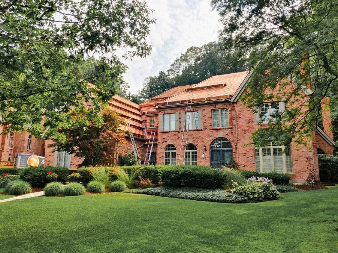 A brick house with a roof under repair, surrounded by a well-maintained lawn and landscaped garden with bushes and flowers.