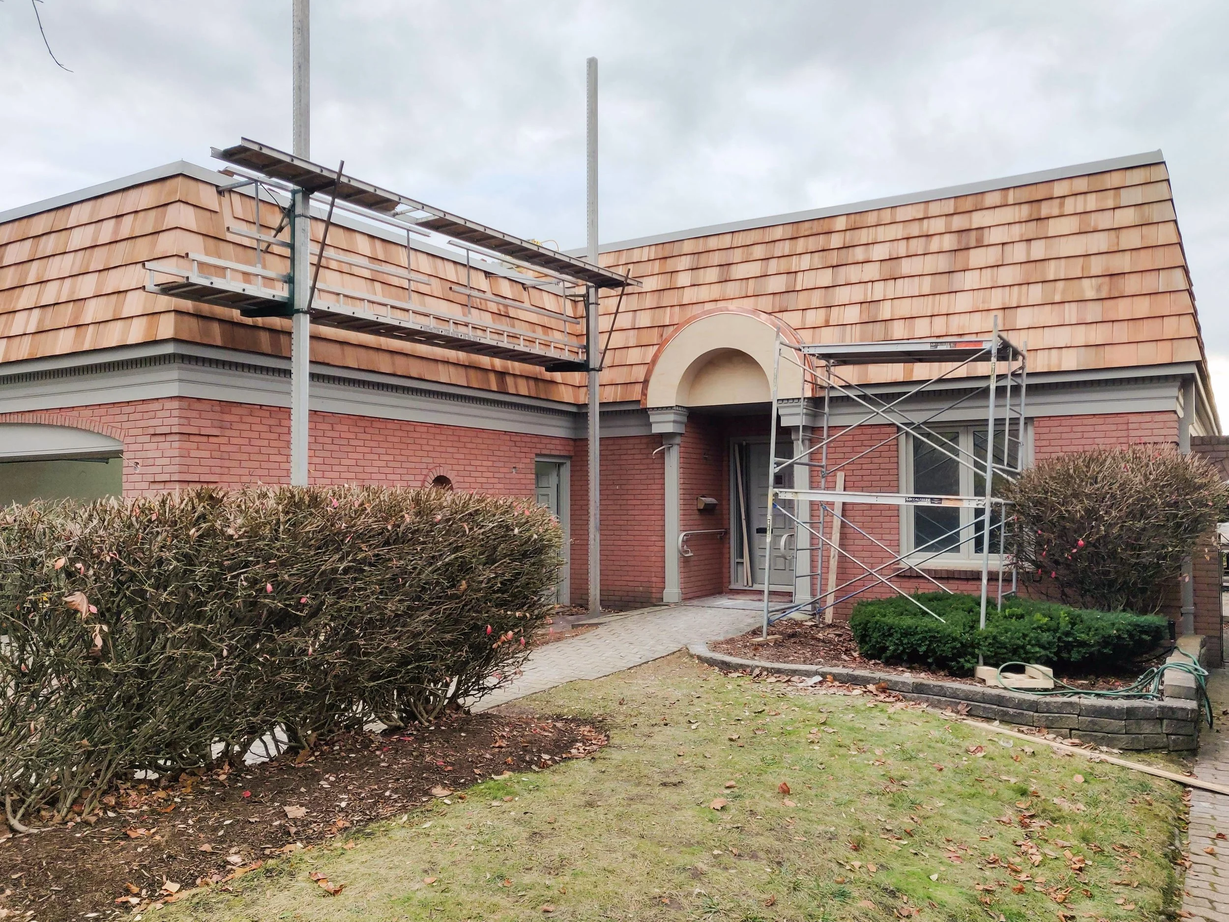 A residential house with scaffolding on the front. The house has brick walls, a shingled roof, and a small front yard with bushes and a paved walkway.