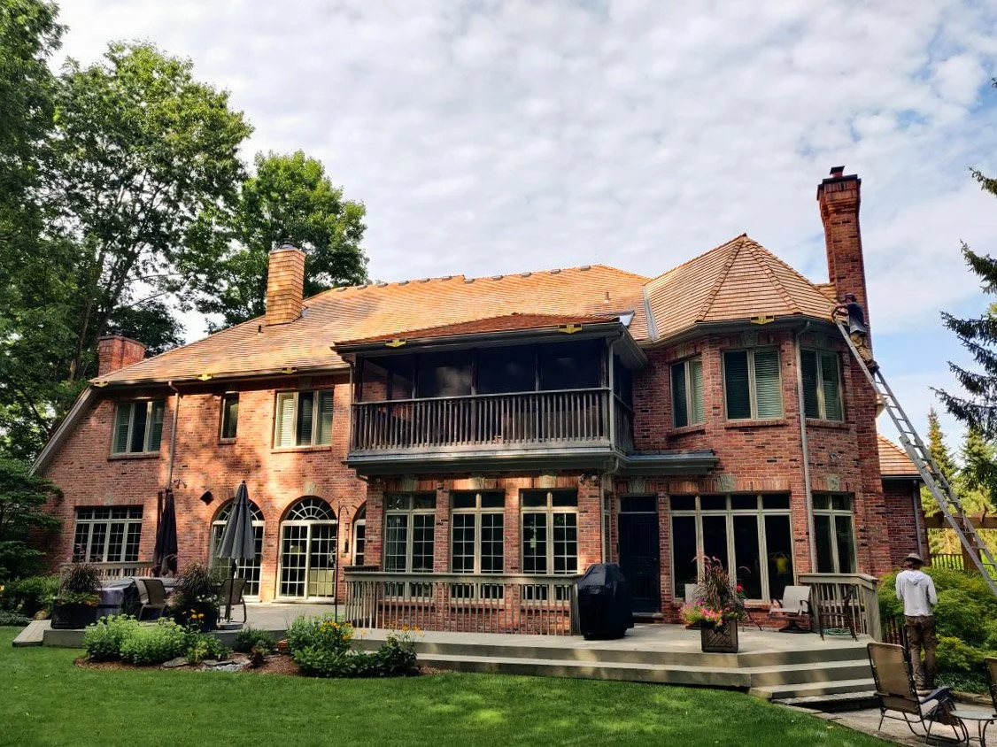 Back view of a large brick house with multiple windows, a screened porch, a balcony, and a tall chimney, surrounded by trees and a well-maintained lawn.
