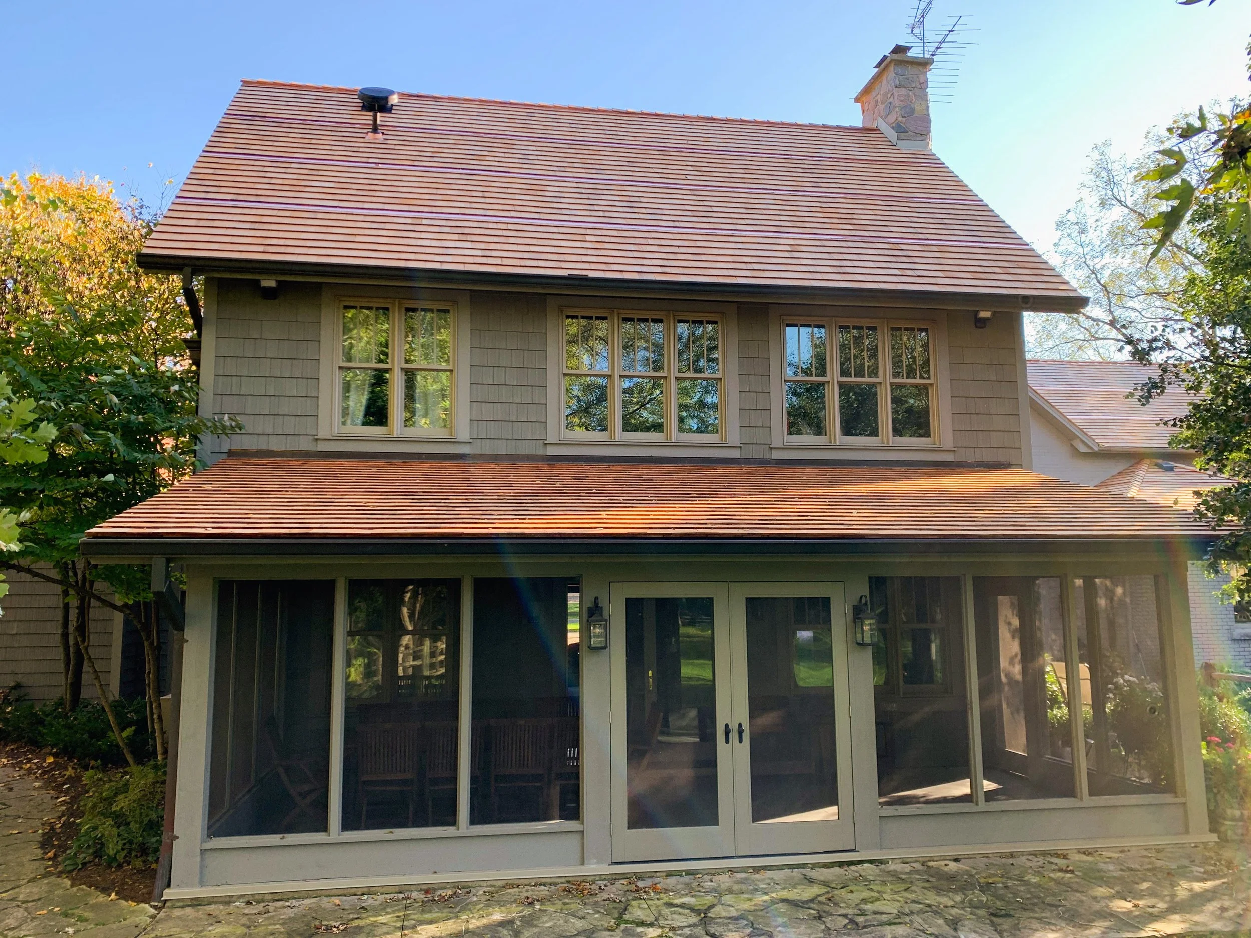 Two-story house with a screened porch, multiple windows, a gabled roof with red shingles, and a stone chimney, surrounded by trees