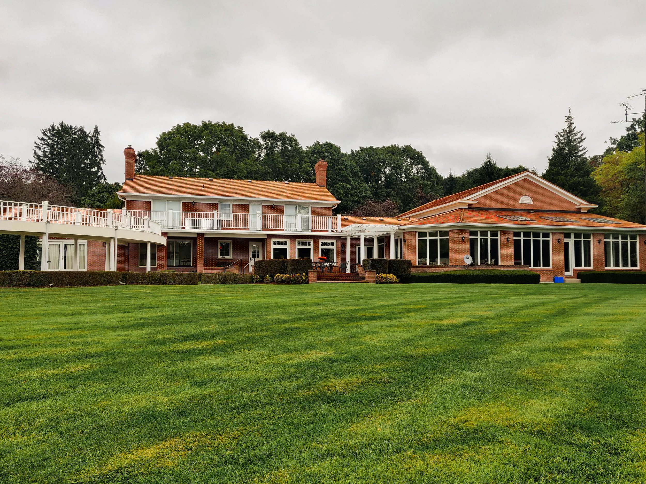 Large brick house with multiple windows, balconies, and a spacious lawn with well-manicured grass, set against a cloudy sky and surrounded by trees.