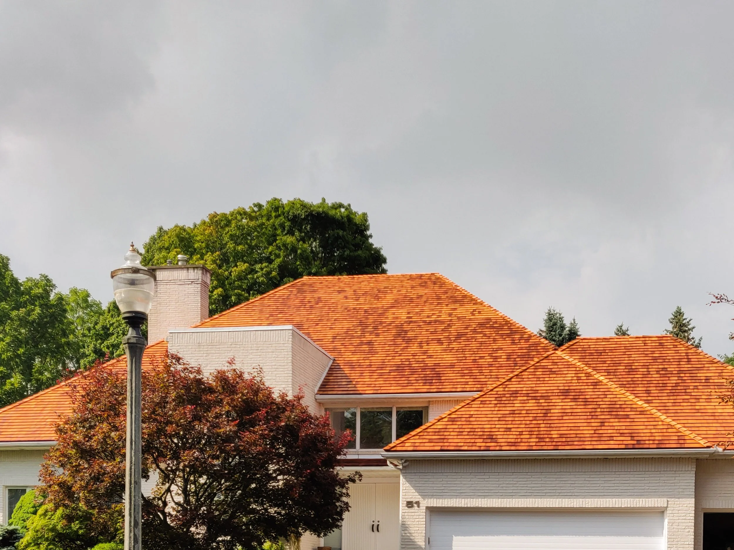 A house with a red tiled roof, white brick exterior, and a large window. There is a streetlamp and trees in the foreground with a cloudy sky above.
