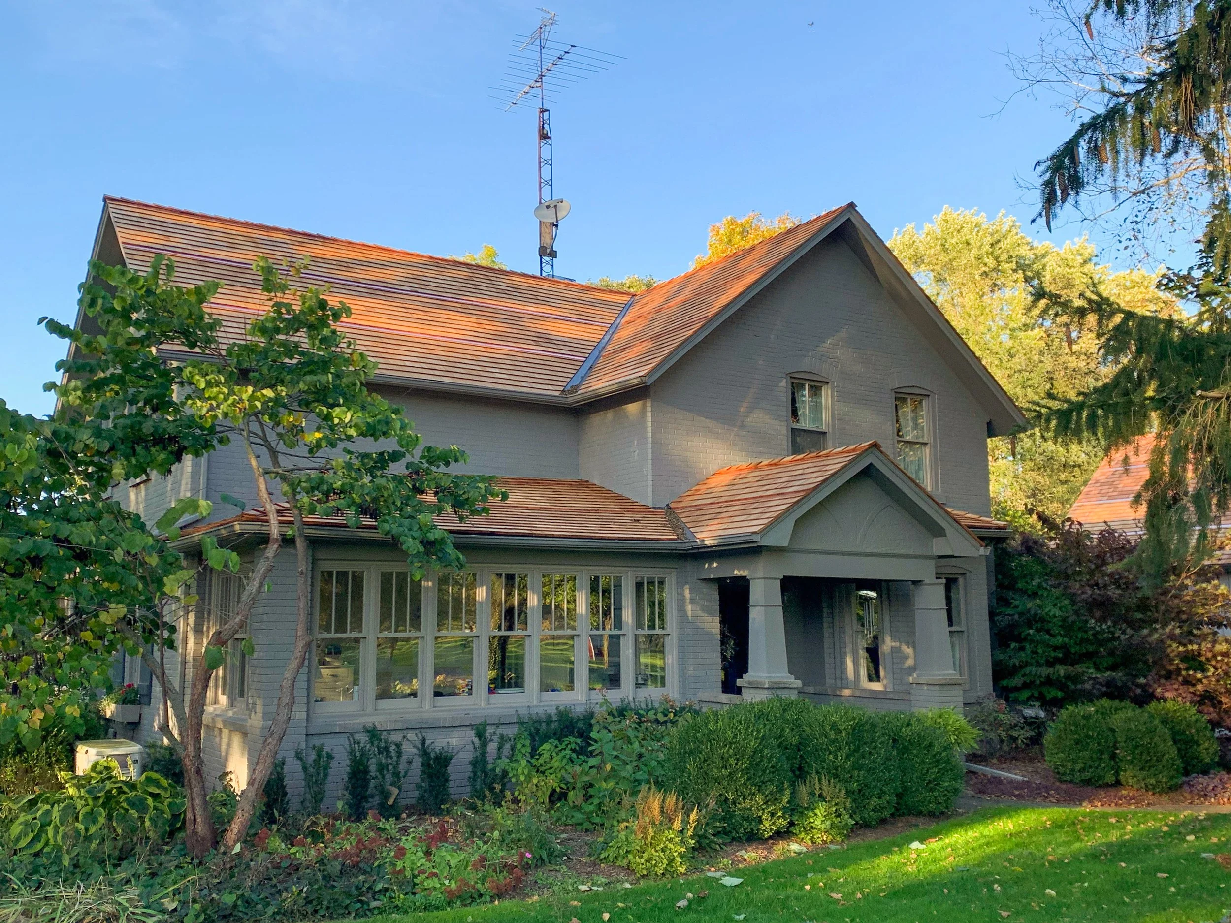 A large two-story house with a gray exterior and a red tiled roof, surrounded by greenery and trees, under a clear blue sky.
