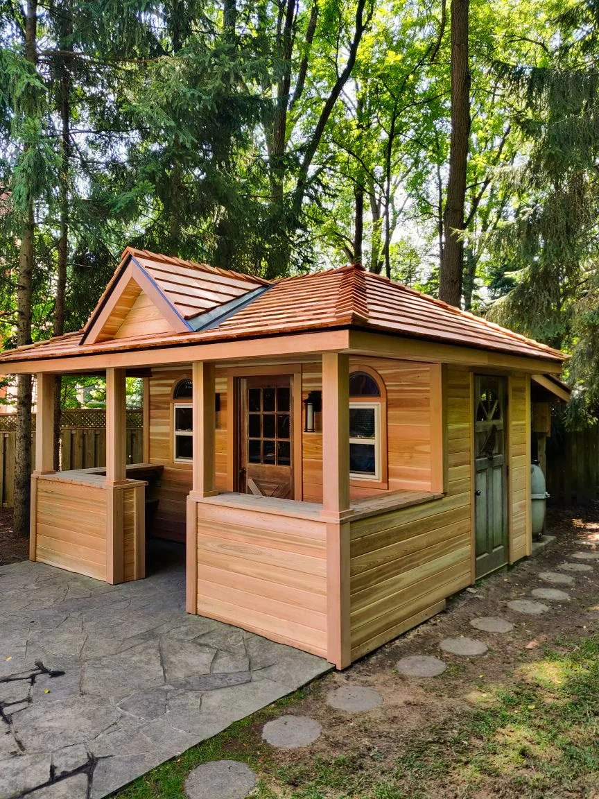 Small wooden playhouse with a reddish-brown tile roof and a small porch, surrounded by trees and a stone pathway.