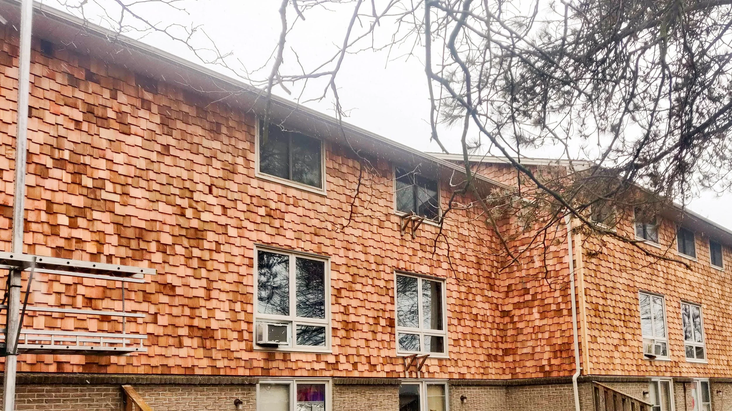 A multi-story brick apartment building with a roof that has new reddish-brown wooden shingles, and several windows with some air conditioning units installed.