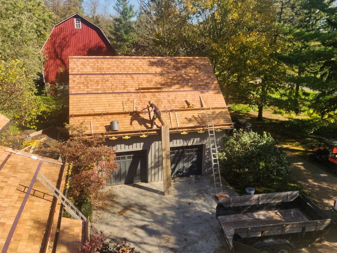 Construction workers installing roofing on a house with a wooden ladder and a truck nearby, surrounded by trees.