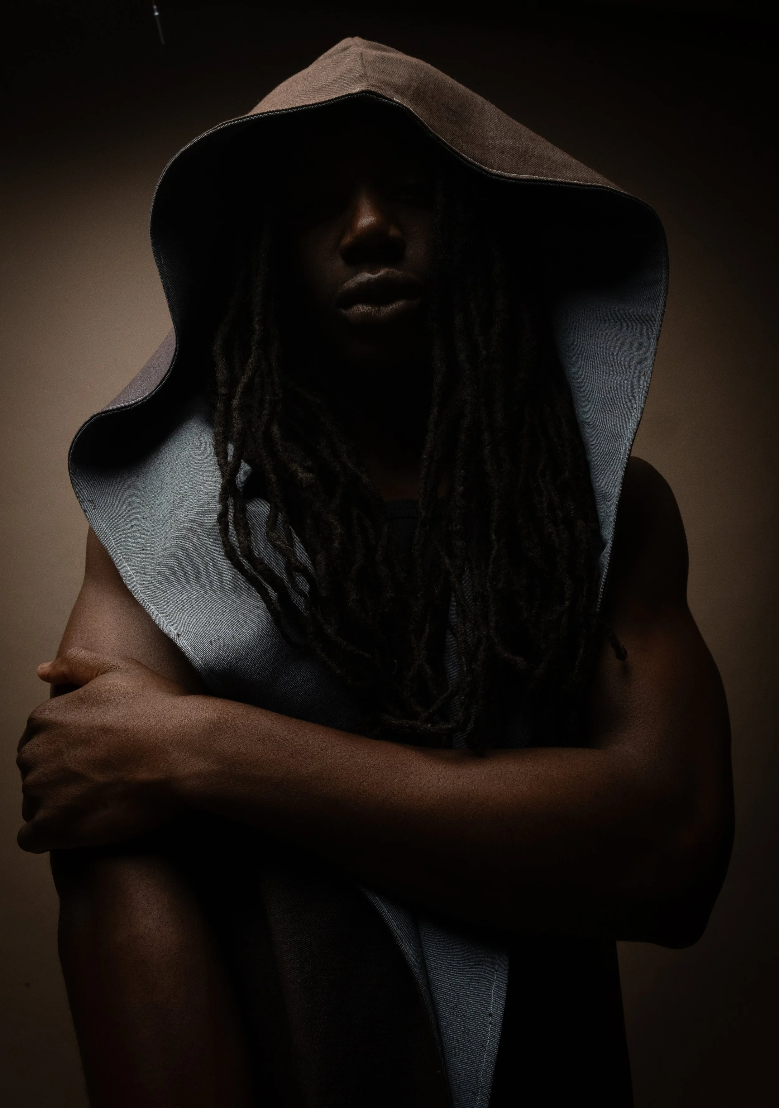 Personne portant une tenue sombre avec un grand chapeau ou capuche, dans une lumière tamisée, couvrant partiellement le visage dans l'ombre.