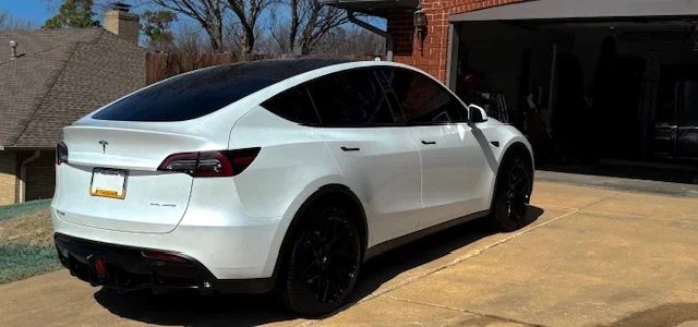 A white Tesla Model X parked in a driveway next to a brick house, with dark tinted windows and black wheels.