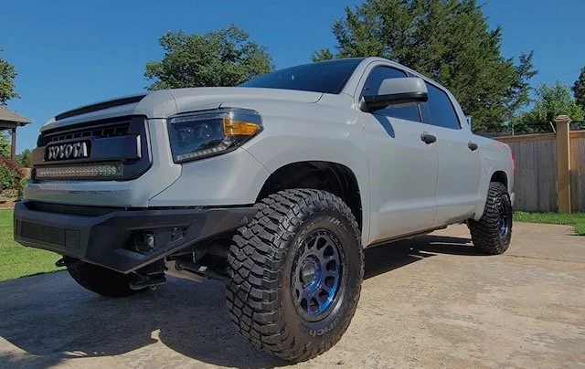 White Toyota pickup truck with large off-road tires parked on a driveway in front of a fence and trees.